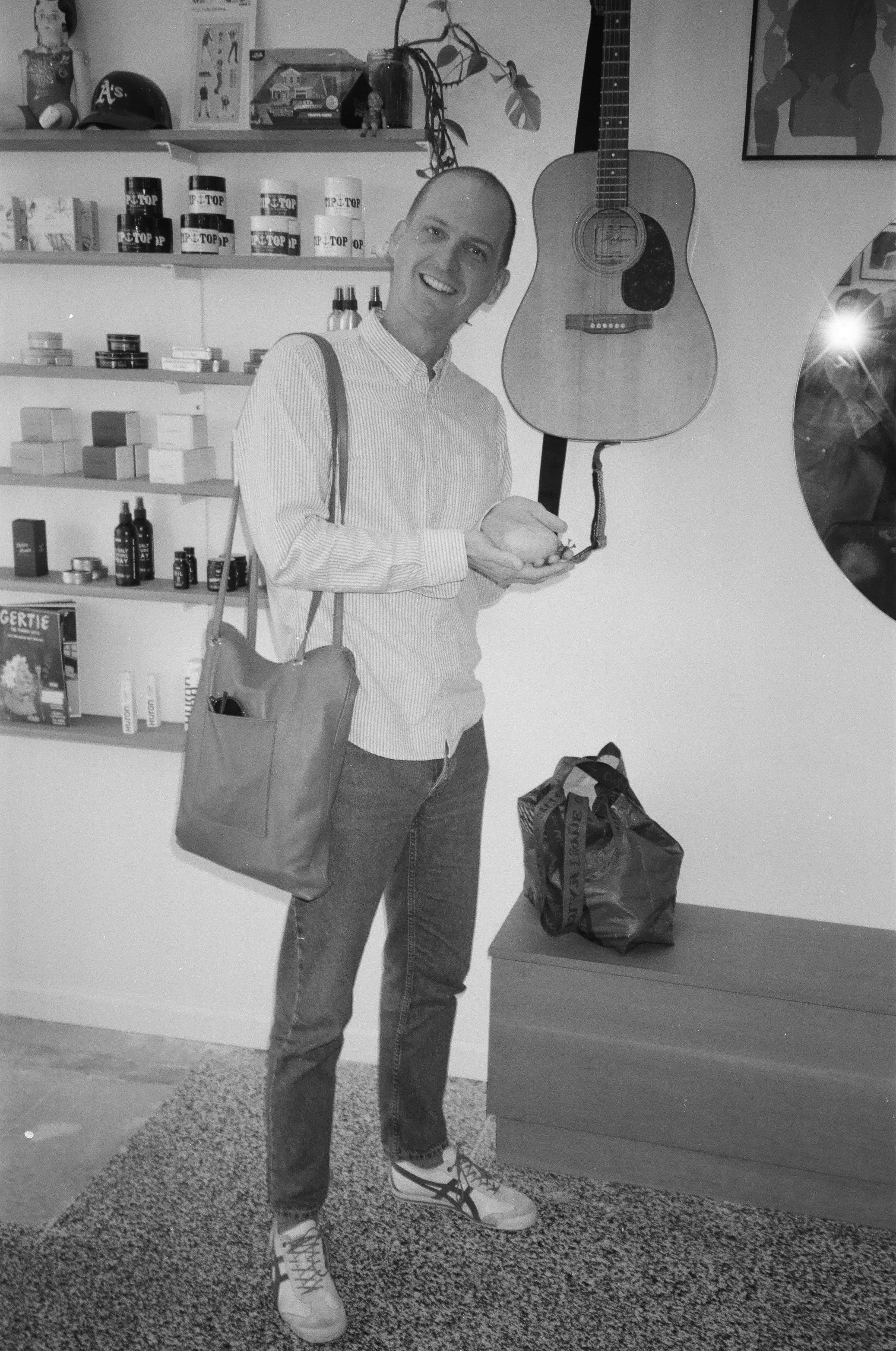 A man smiling and standing indoors near shelves filled with various products, with a guitar hanging on the wall behind him, a small dark bag on a low table, and a pile of items on the floor.