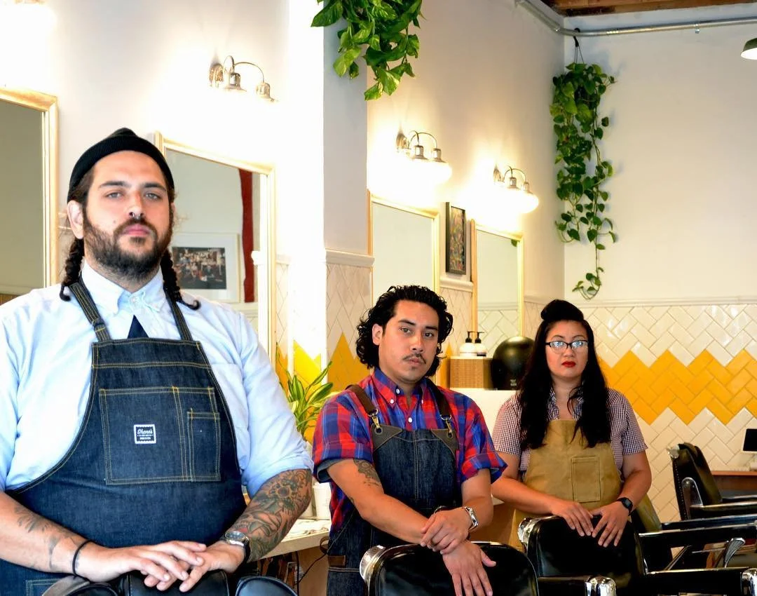 Three staff members in a restaurant with white and yellow decor, wearing aprons, standing behind chairs.