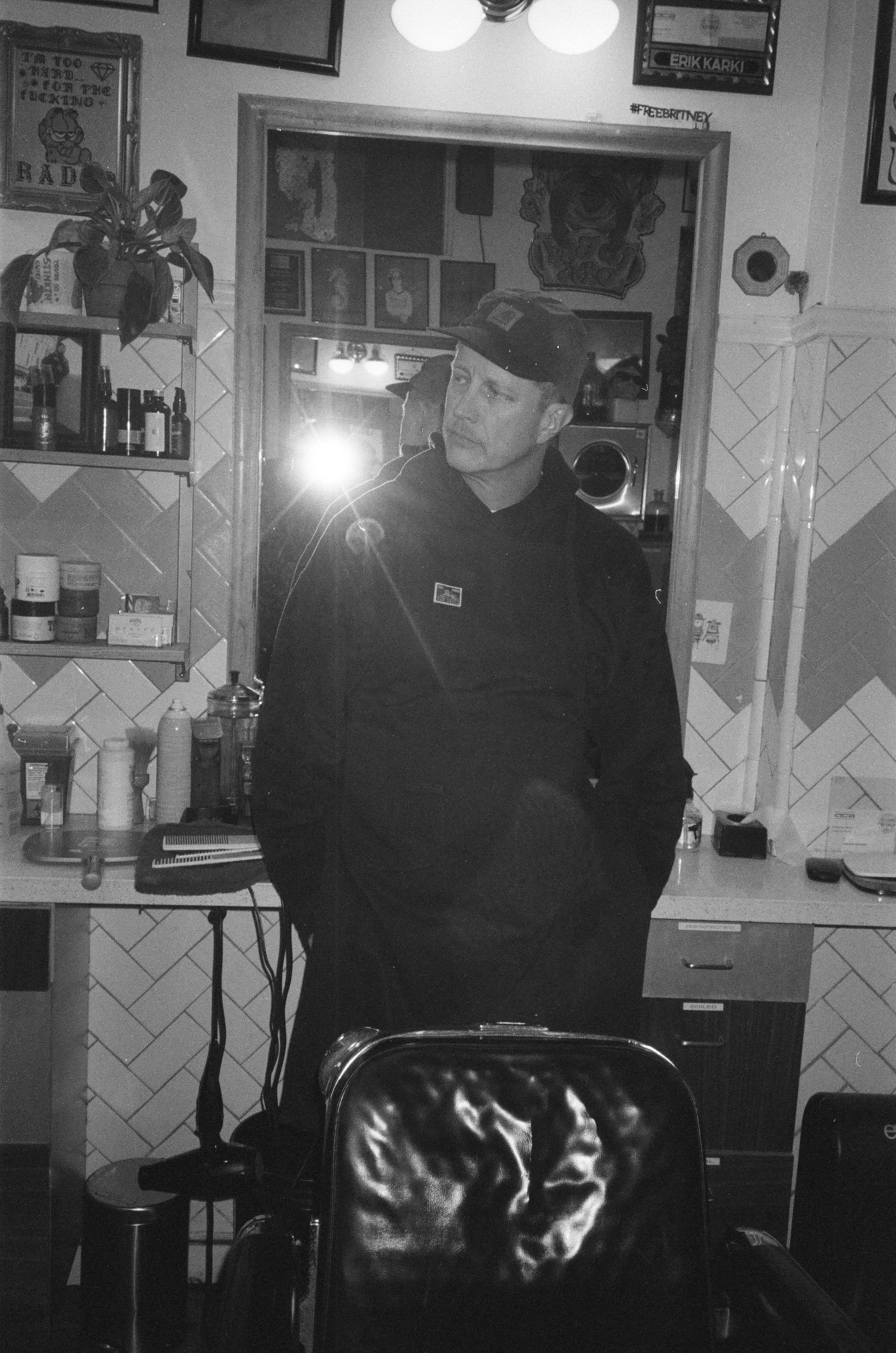 A man wearing a black jacket and a baseball cap standing in a room with patterned tiles on the wall. Behind him, there is a mirror, shelves with bottles, framed pictures, and artwork, with another person visible in the reflection.