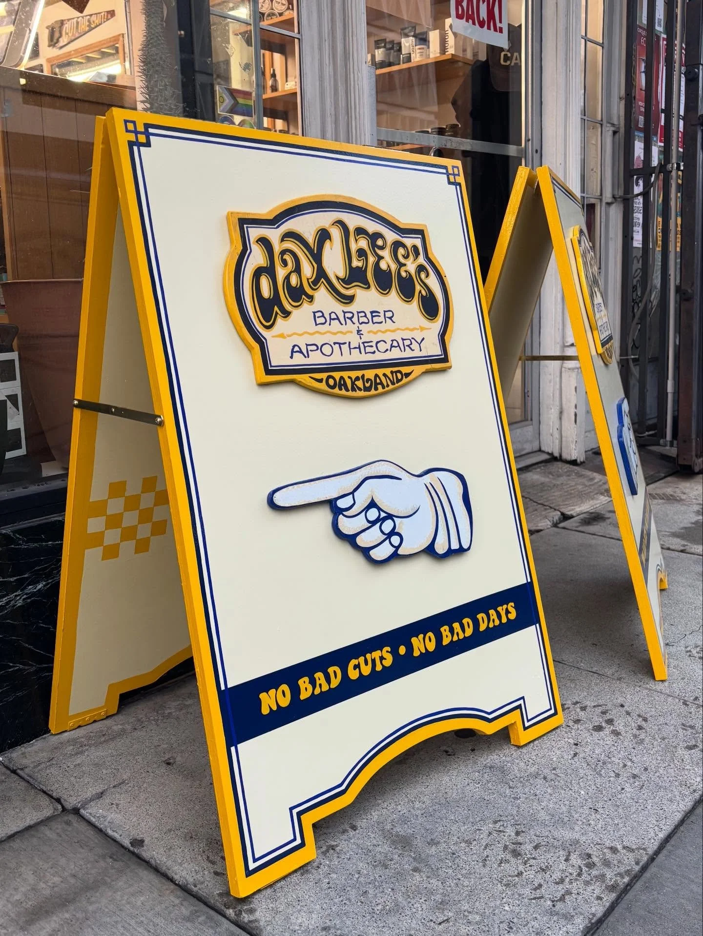 A sandwich board sign outside a business, advertising a barber and apothecary shop in Oakland. The sign features the business logo and a blue and yellow color scheme, along with a graphic of a pointing hand and the message: 'No bad cuts - No bad days