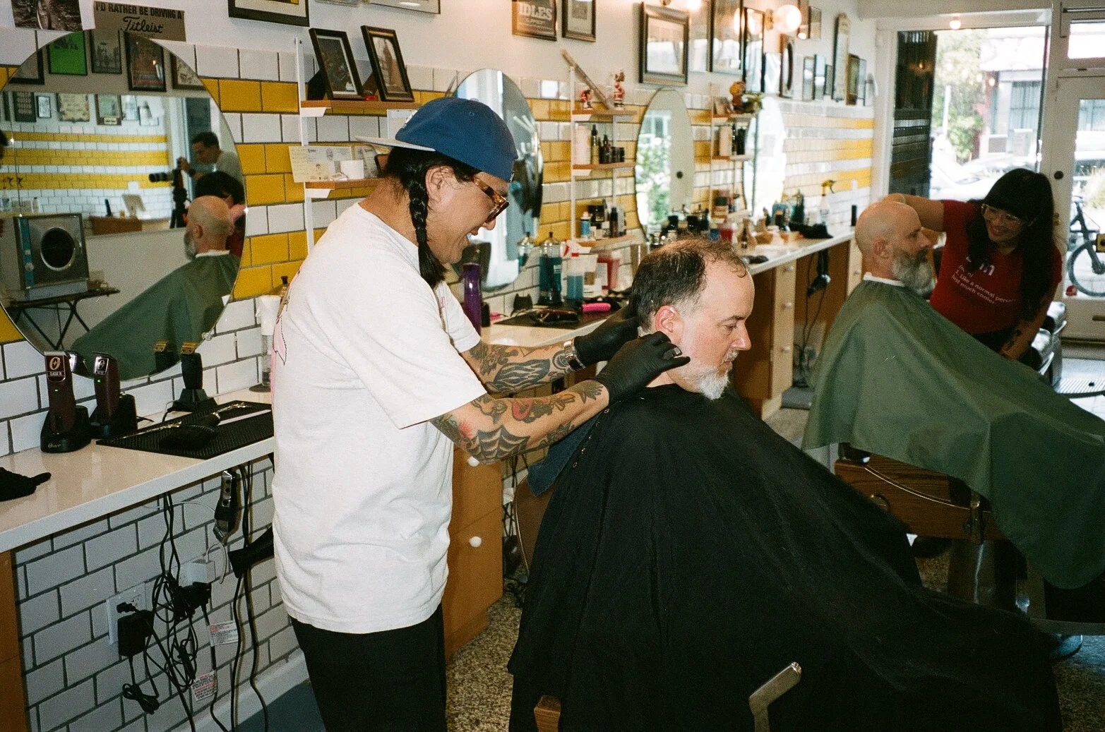Barber shop with two hairstylists cutting men's hair. One stylist has tattoos and is smiling while working on a client; the other stylist is also styling another customer. The shop has a modern decor with mirrors, framed pictures, and various hair pr
