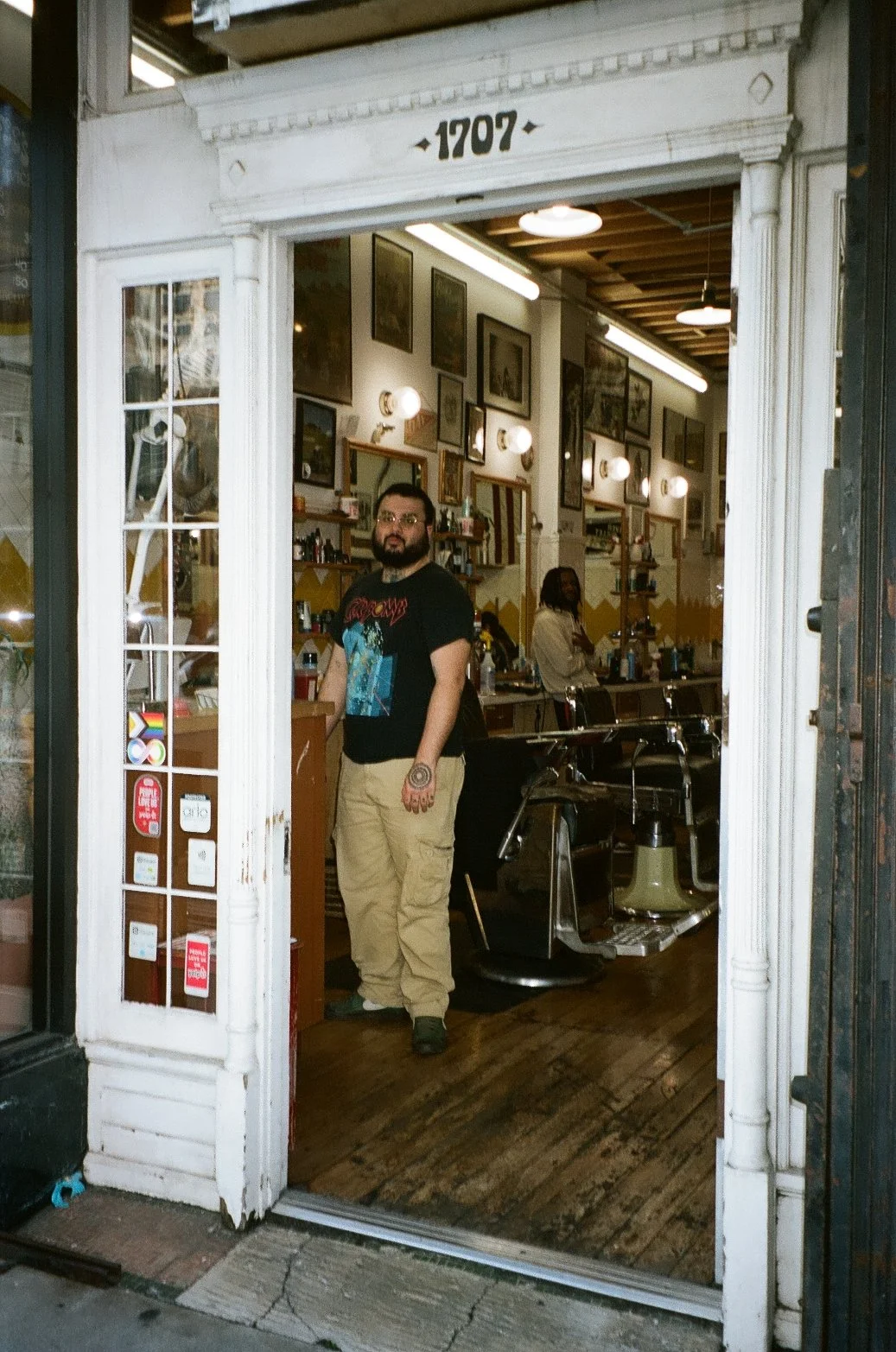 View of a barber shop with a man standing inside, facing forward, wearing a black t-shirt with colorful print and khaki pants. The shop has framed pictures on the walls, barber chairs, and supplies on shelves. The shop is lit with warm lighting and h