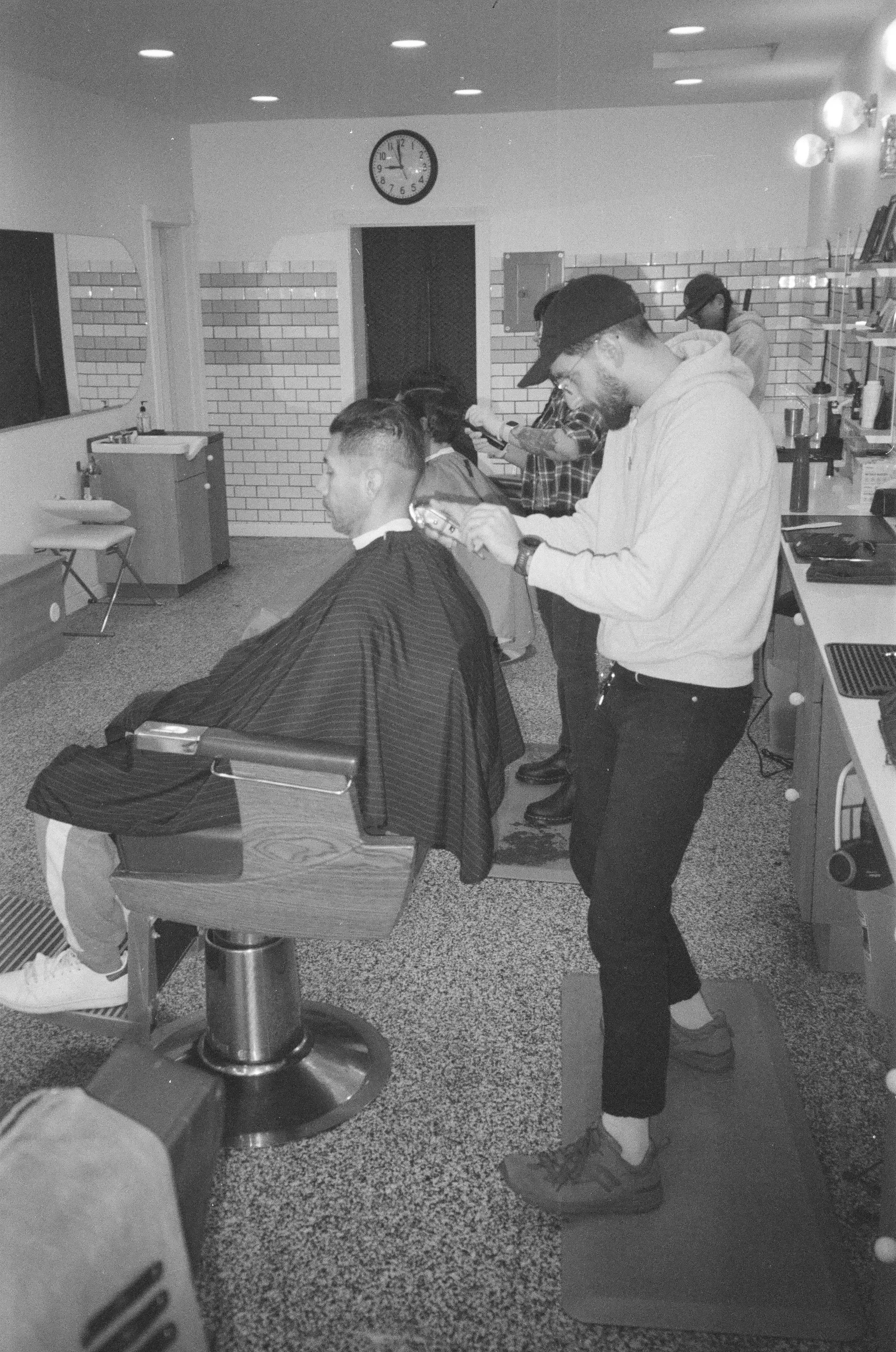 Black and white photo of a barbershop with three men getting haircuts. One man is seated in a barber chair, while another barber trims his hair with clippers. The other barbershop worker is in the background. The room has exposed brick walls, a wall 