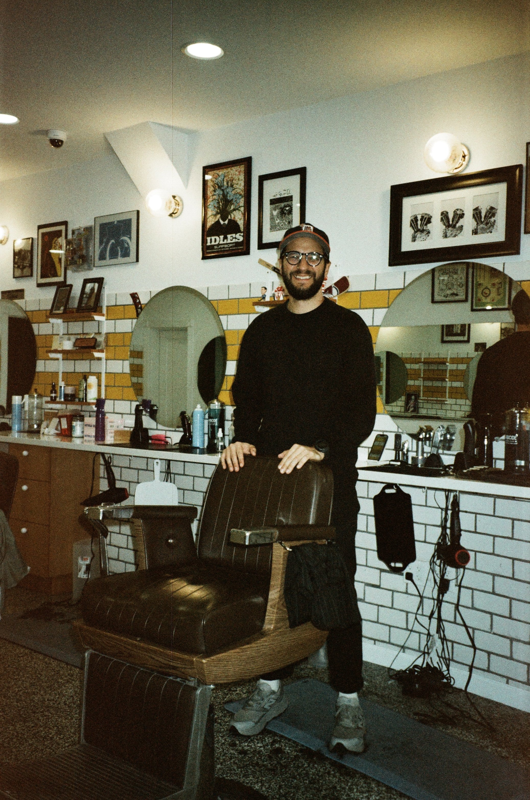A man with glasses and a beard smiling, standing in a barbershop, with various hair and grooming products on the counter and a vintage barber chair in front of him. Behind him are framed pictures and artwork on a white brick wall, illuminated by wall