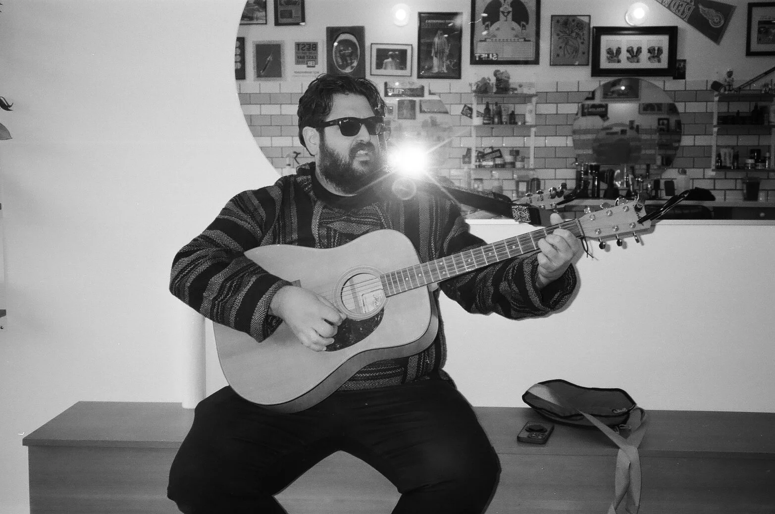 A man with dark hair, beard, and sunglasses playing an acoustic guitar. He is sitting on a bench in a room decorated with framed pictures and a brick wall.