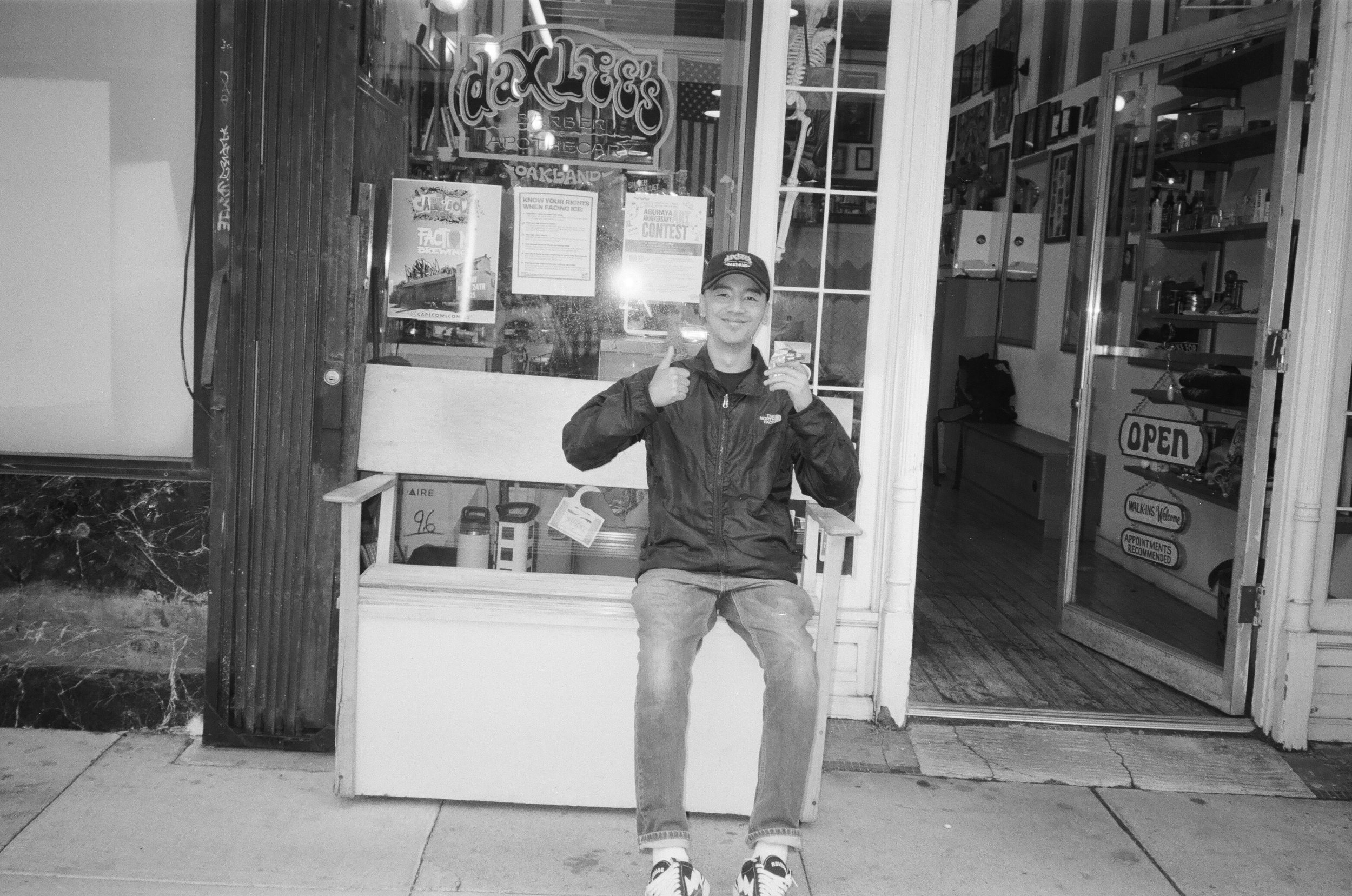 A young man wearing a cap and jacket sitting on a bench outside a shop, smiling and giving a thumbs-up. The shop has signs and posters on the windows, with shelves visible inside.