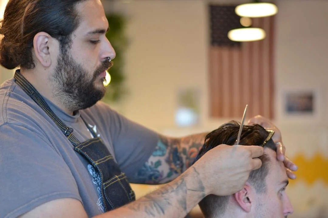 Barber cutting a client's hair in a barbershop with American flag and curtains in the background.