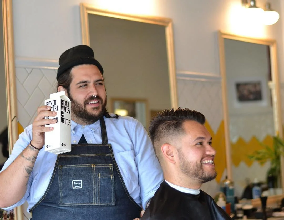 Barber is cutting a man's hair and both are smiling in a barber shop with mirrors in the background.