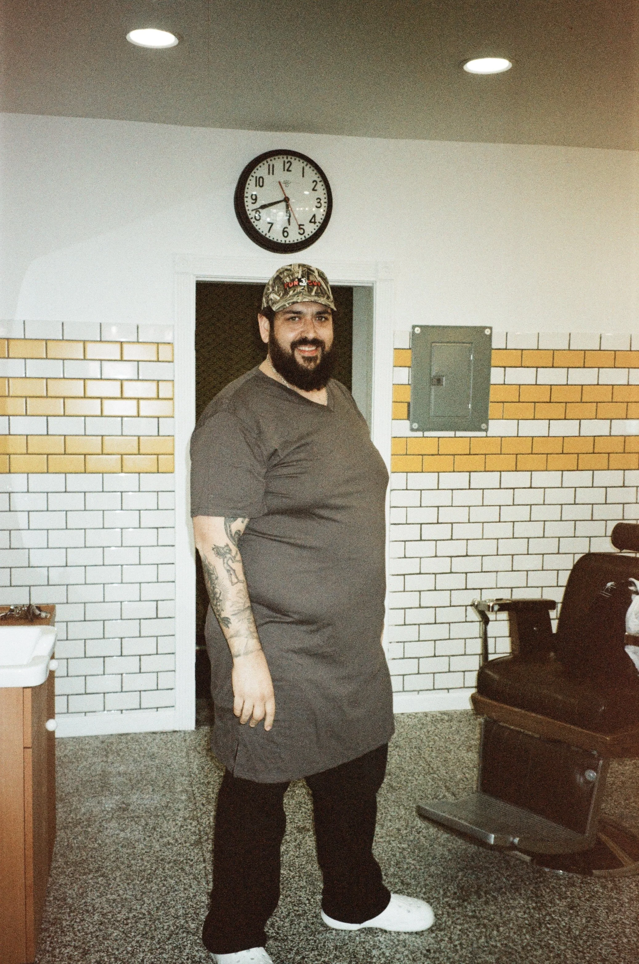 A man with a beard and tattoos on his left arm, wearing a camouflage cap, gray t-shirt, black pants, and white sneakers, is standing indoors with a brick wall background, a wall clock showing 8:17, and a black chair.