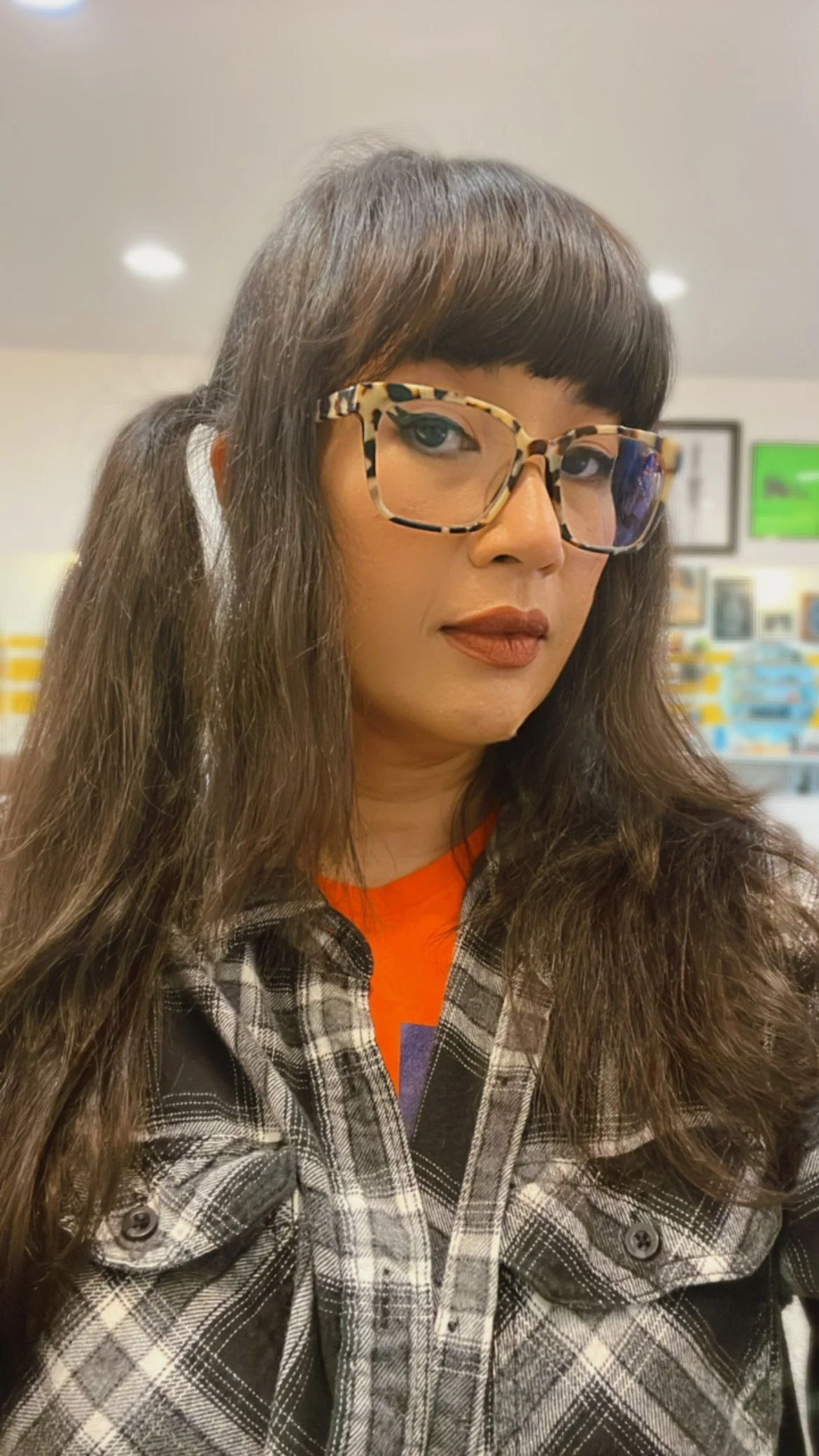 A woman with brown hair and bangs wearing leopard print glasses, an orange shirt, and a black and white plaid jacket, standing indoors with framed pictures on the wall behind her.