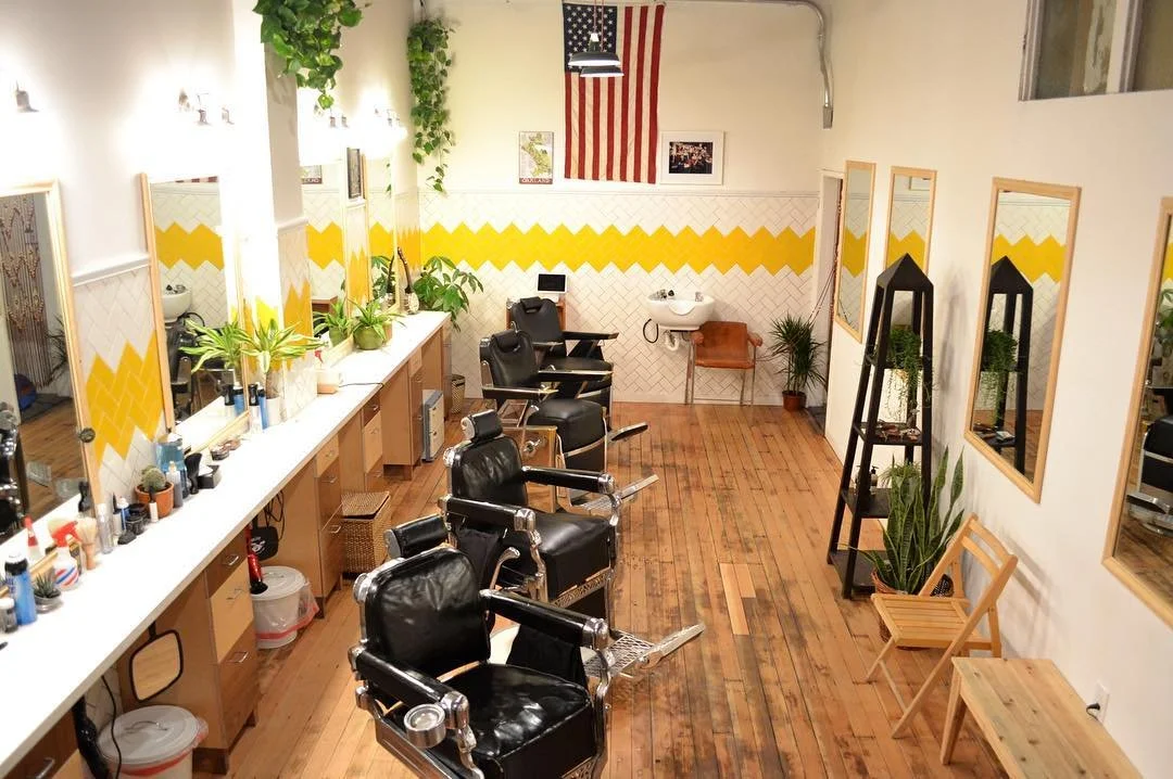 Empty barbershop with four black chairs, a long mirror wall, plants, and an American flag on the wall.