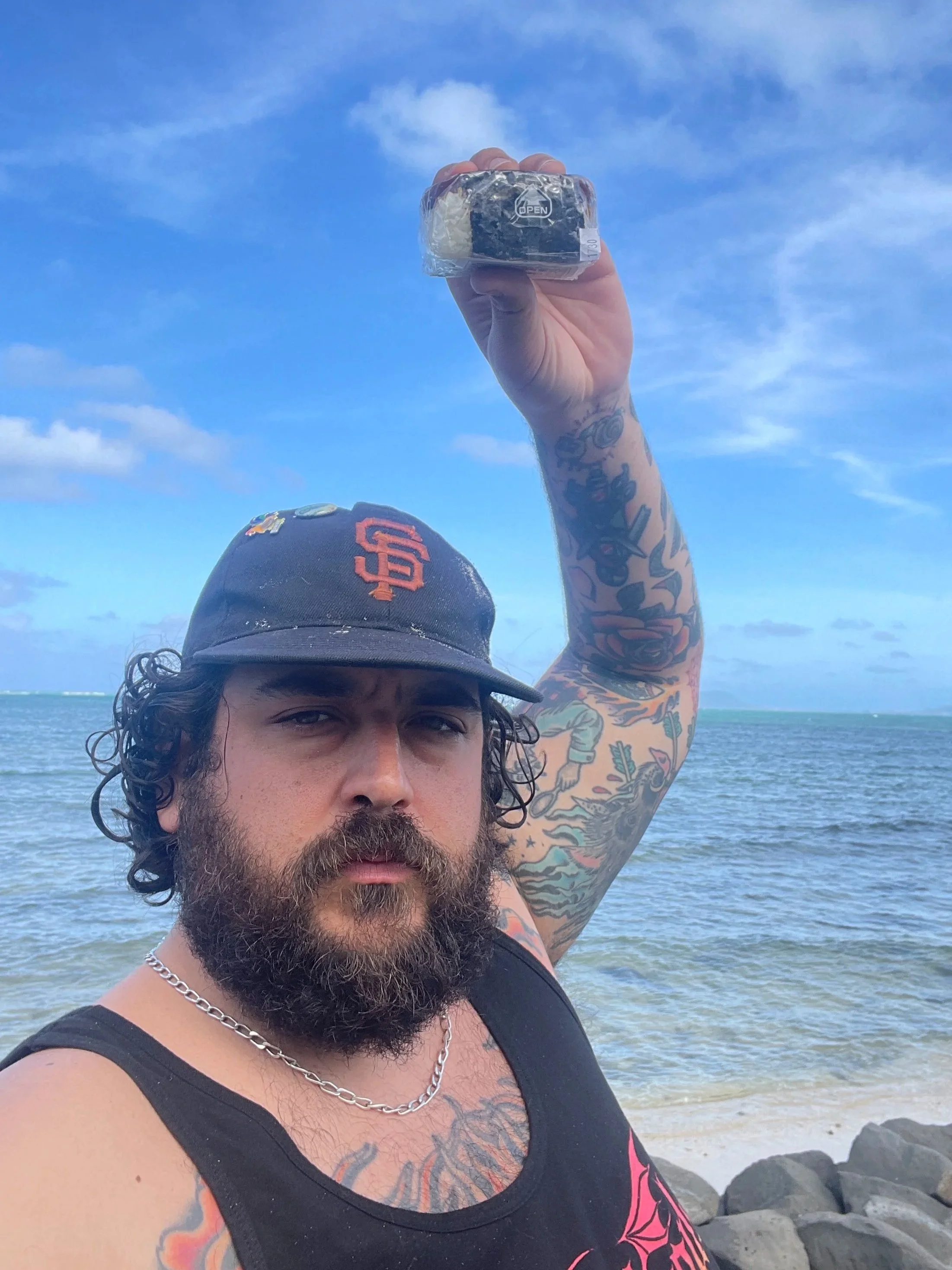 A man with tattoos, a beard, and a chain necklace takes a selfie at the beach, wearing a black sleeveless shirt and a black baseball cap with an orange logo, with the ocean and rocks in the background.