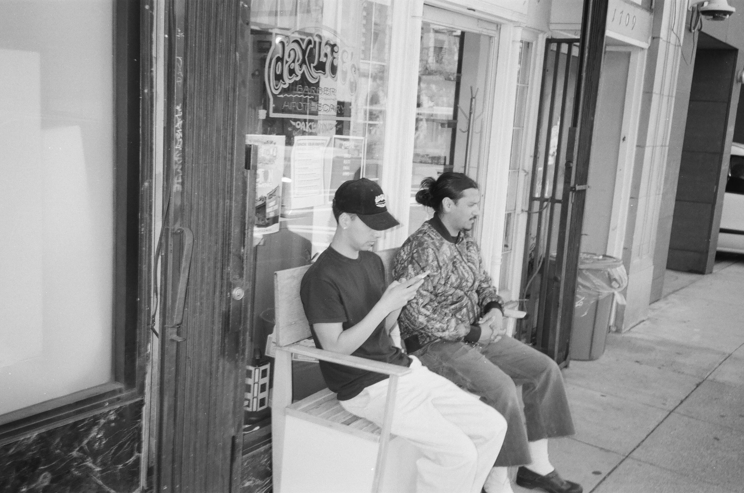 Two people sitting on a wooden bench in front of a storefront, one is a young person with a cap and the other is an older woman with a bun, both looking at their phones. The storefront has large windows and a sign, and there is a vehicle partially vi
