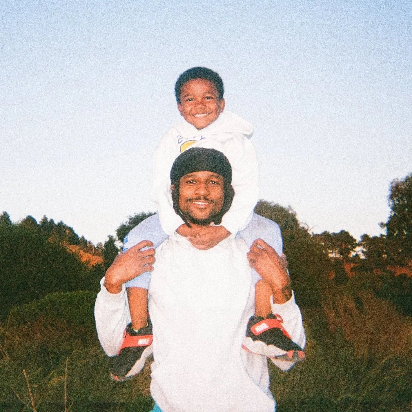 Two smiling males outdoors, one carrying the other on his shoulders, with trees and a clear sky in the background.