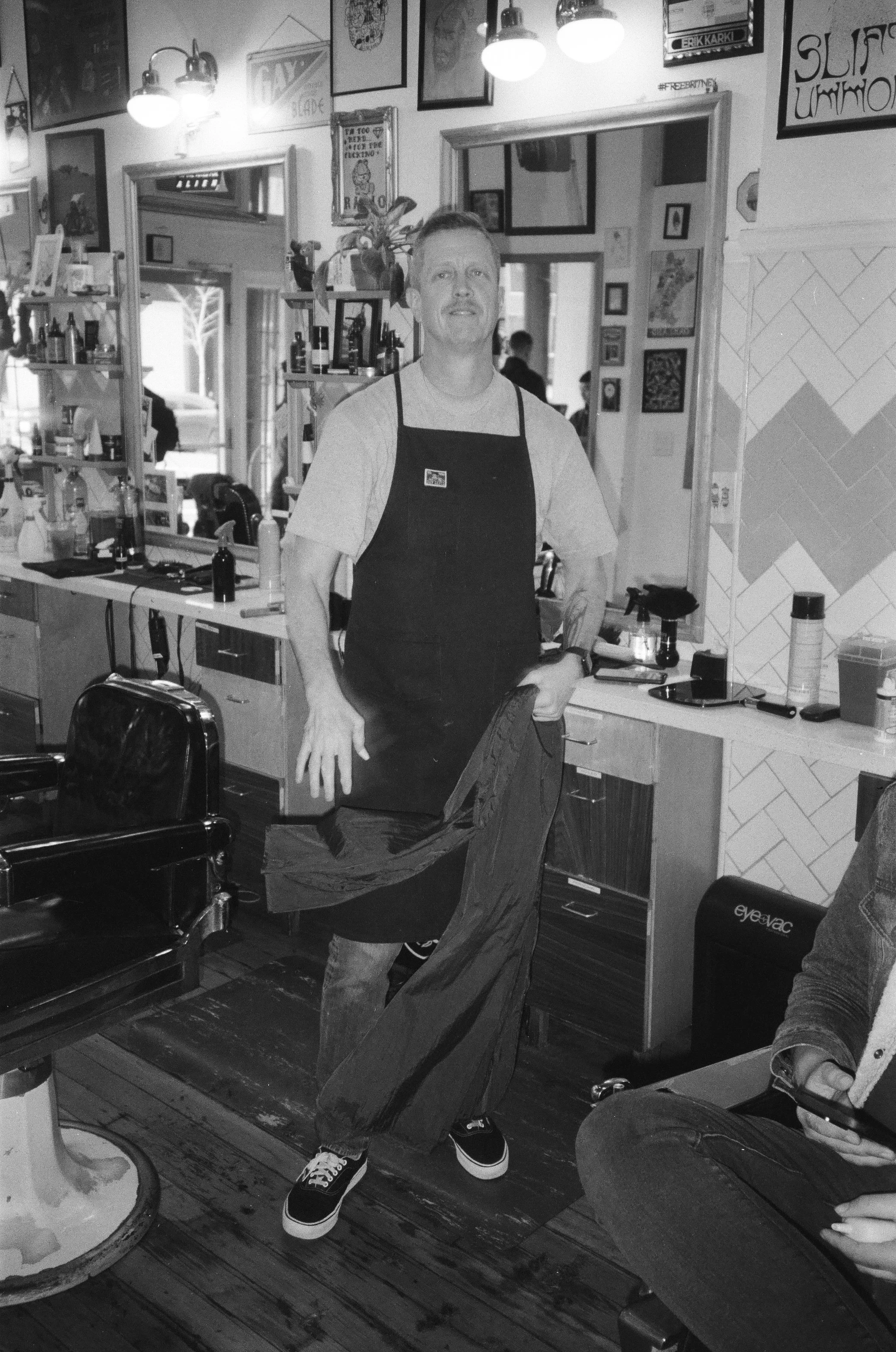 A man wearing a black apron, t-shirt, and sneakers standing in a restaurant or cafe, with surrounding posters and pictures on the wall, and a mirror reflecting other people and objects in the background.