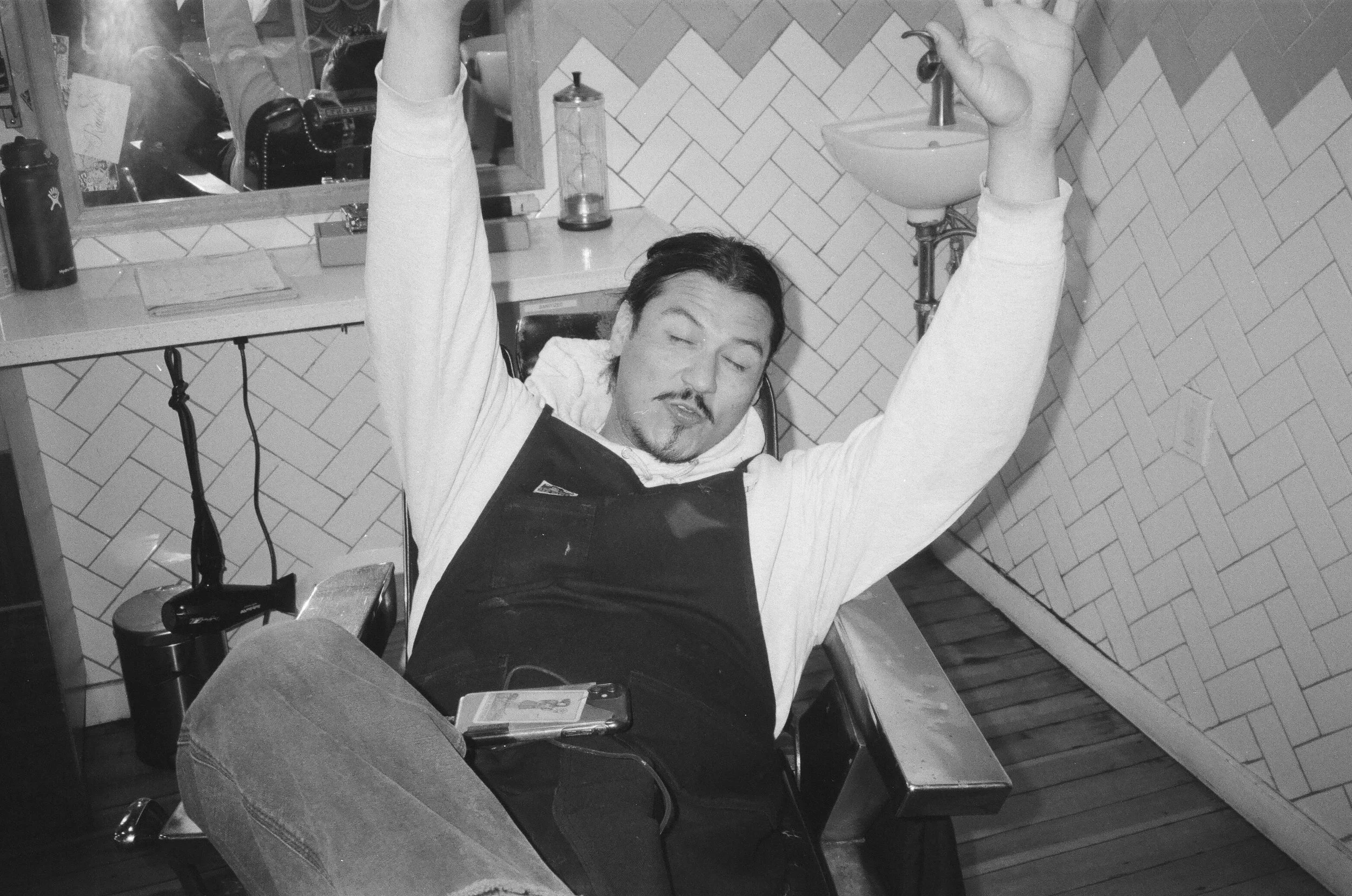 A man with dark hair, facial hair, and closed eyes is reclining in a barber chair with his arms raised above his head. He is wearing a white shirt and a dark apron. The background shows a mirror, a countertop with various barber tools, and a tiled wa