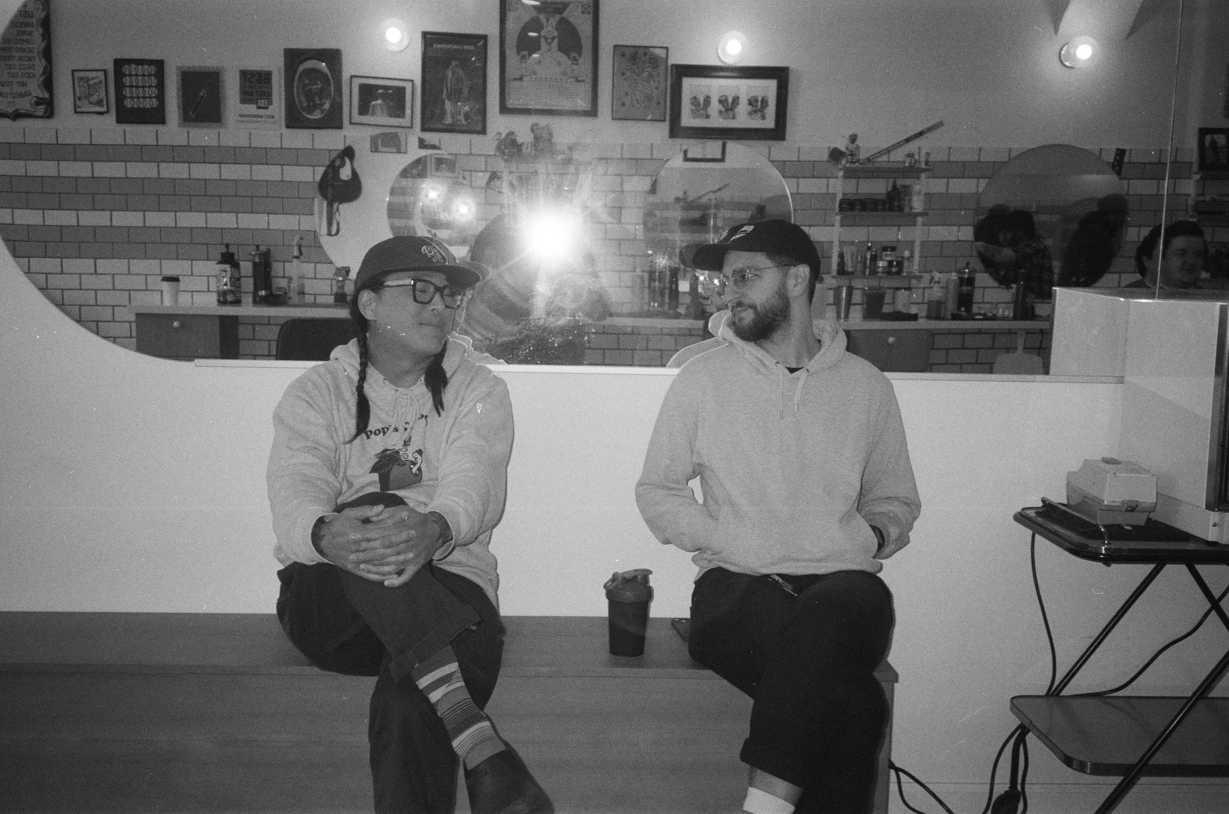 Two young adults sitting at a table in a room with the wall behind them decorated with framed pictures and artworks. The person on the left wears glasses, a baseball cap, and a Hoodie with a graphic, while the person on the right wears a hoodie and a