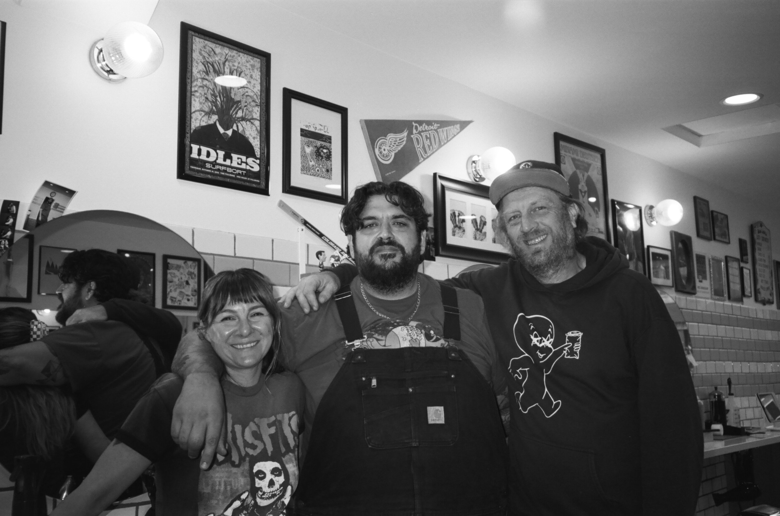 Three people standing together in a restaurant or cafe, smiling. Behind them is a wall decorated with framed posters and artworks, including a Detroit Red Wings banner. The scene is lively with other patrons visible in the background.