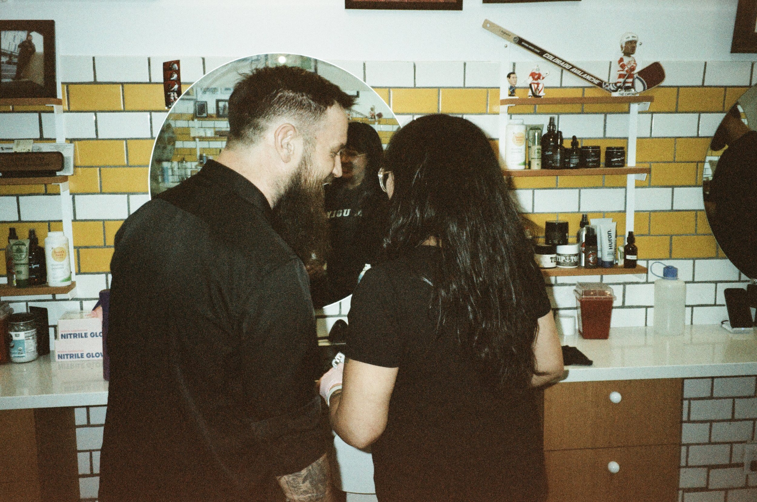 Two people with dark hair and a beard are standing close together, possibly engaging in a conversation. They are inside a room with yellow and white tiled walls, and shelves holding various bottles and products. The man has short hair and a beard, wh