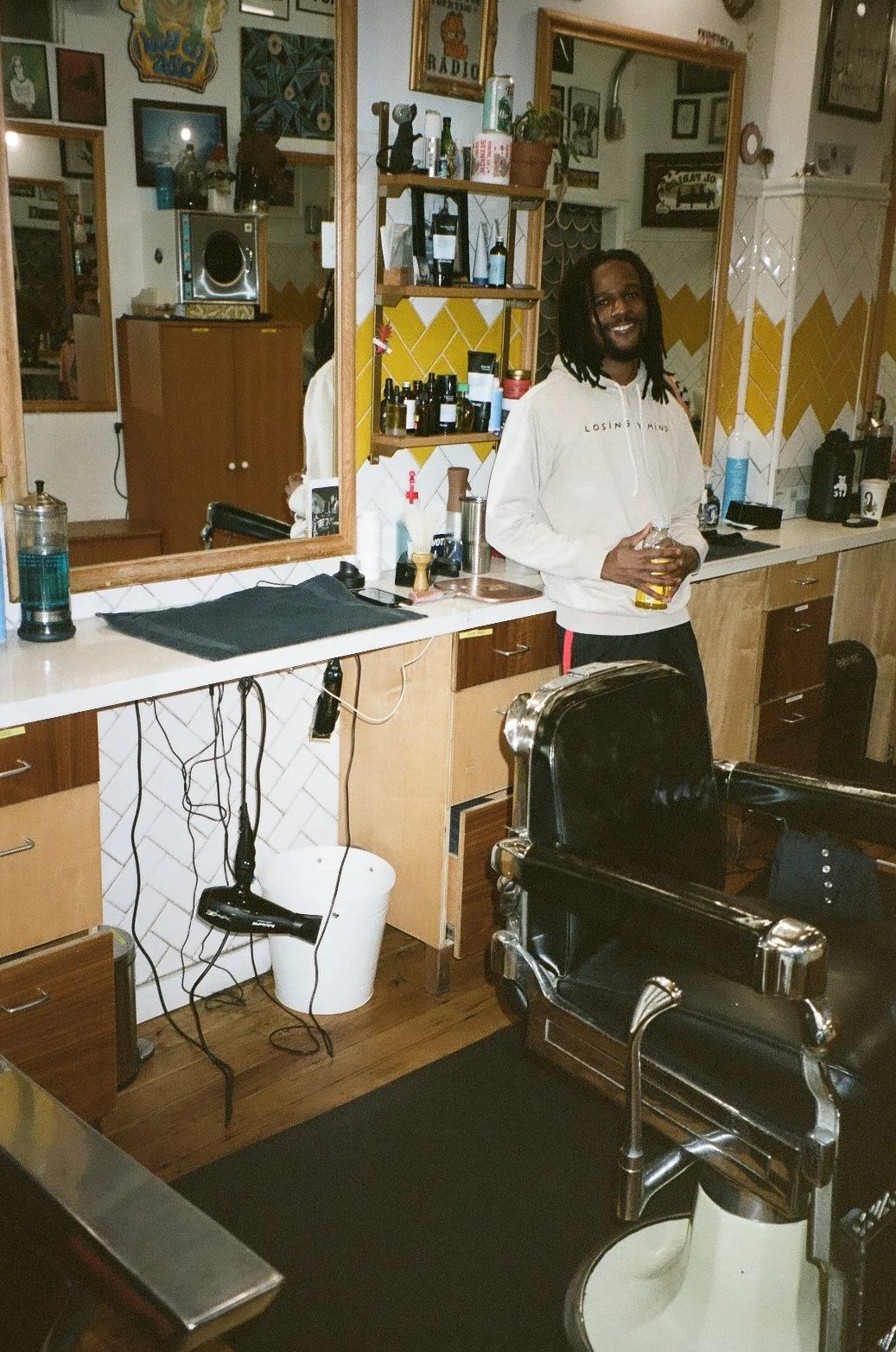 A man with dreadlocks and a light-colored hoodie stands in a vintage barbershop or salon, holding a glass of beer, smiling. The room has mirrors, shelves with bottles, a barber chair, and various hair and grooming tools.