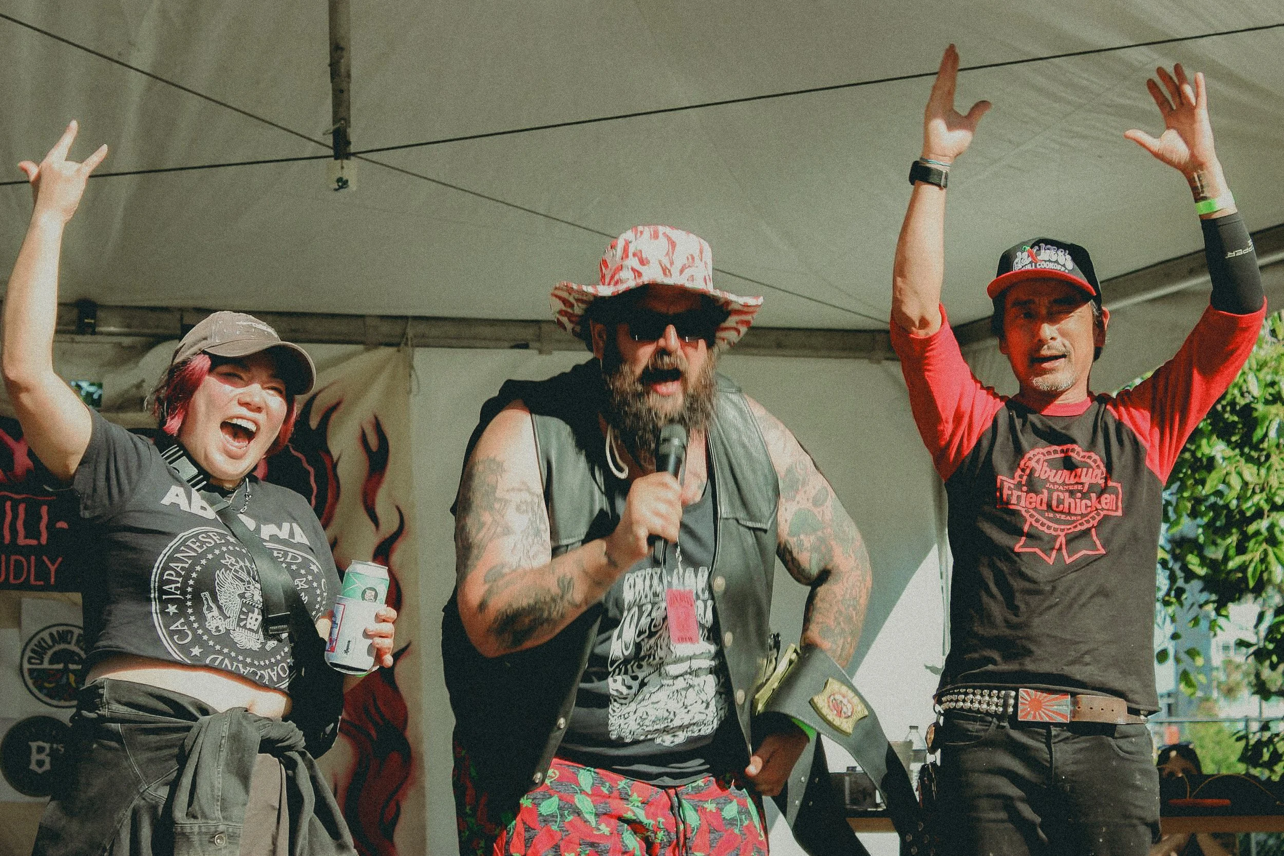Three people on a stage celebrating; a woman on the left with a gray cap and a black T-shirt, a man in the middle with a red and white hat, sunglasses, tattoos, and a black vest, and a man on the right in a red and black T-shirt and a black cap.
