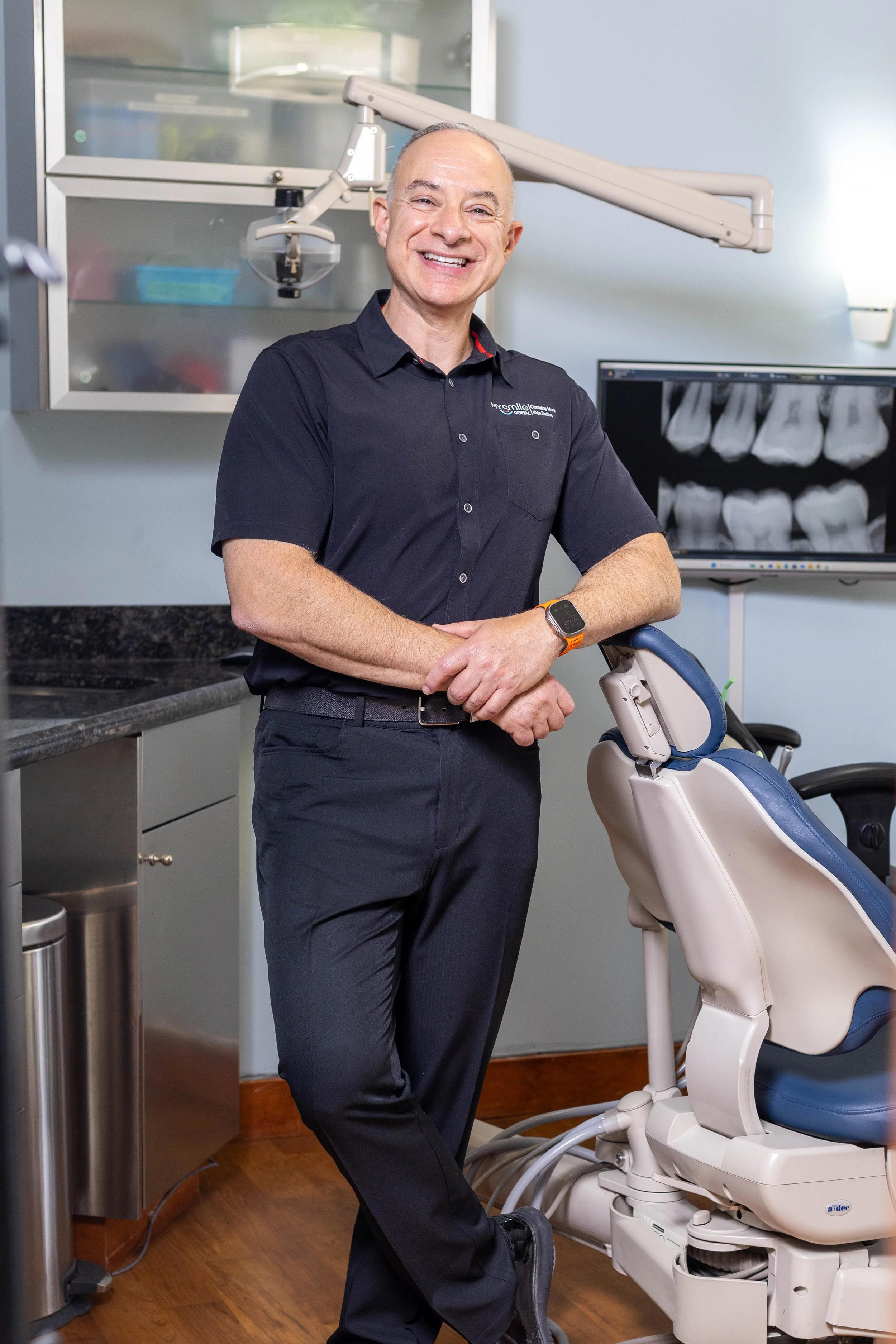 A smiling man in a black shirt and pants standing next to a dental chair in a dental office with dental X-ray images on a monitor in the background.