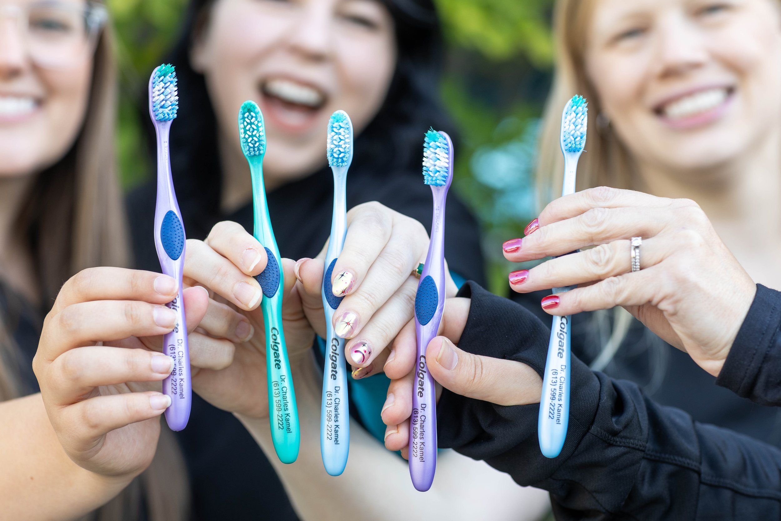 Three women holding colorful toothbrushes outdoors, smiling and showing teeth, with greenery in the background.