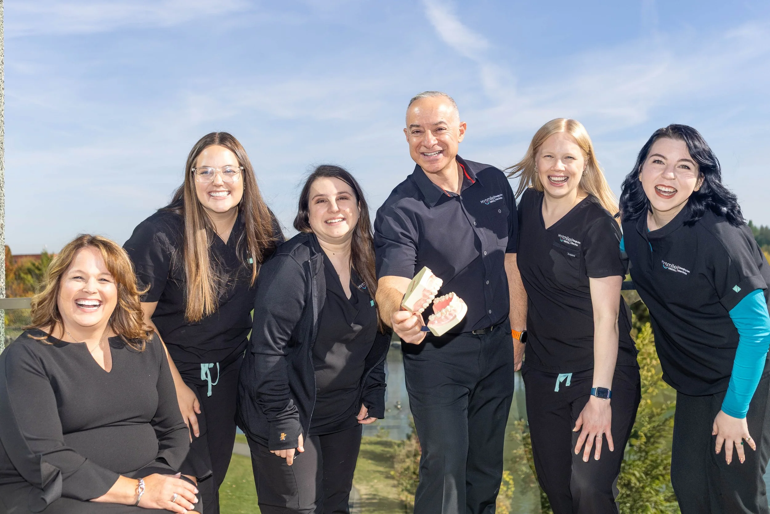 Group of six women and one man smiling outdoors, with the man holding a dental model, under a blue sky with some clouds.