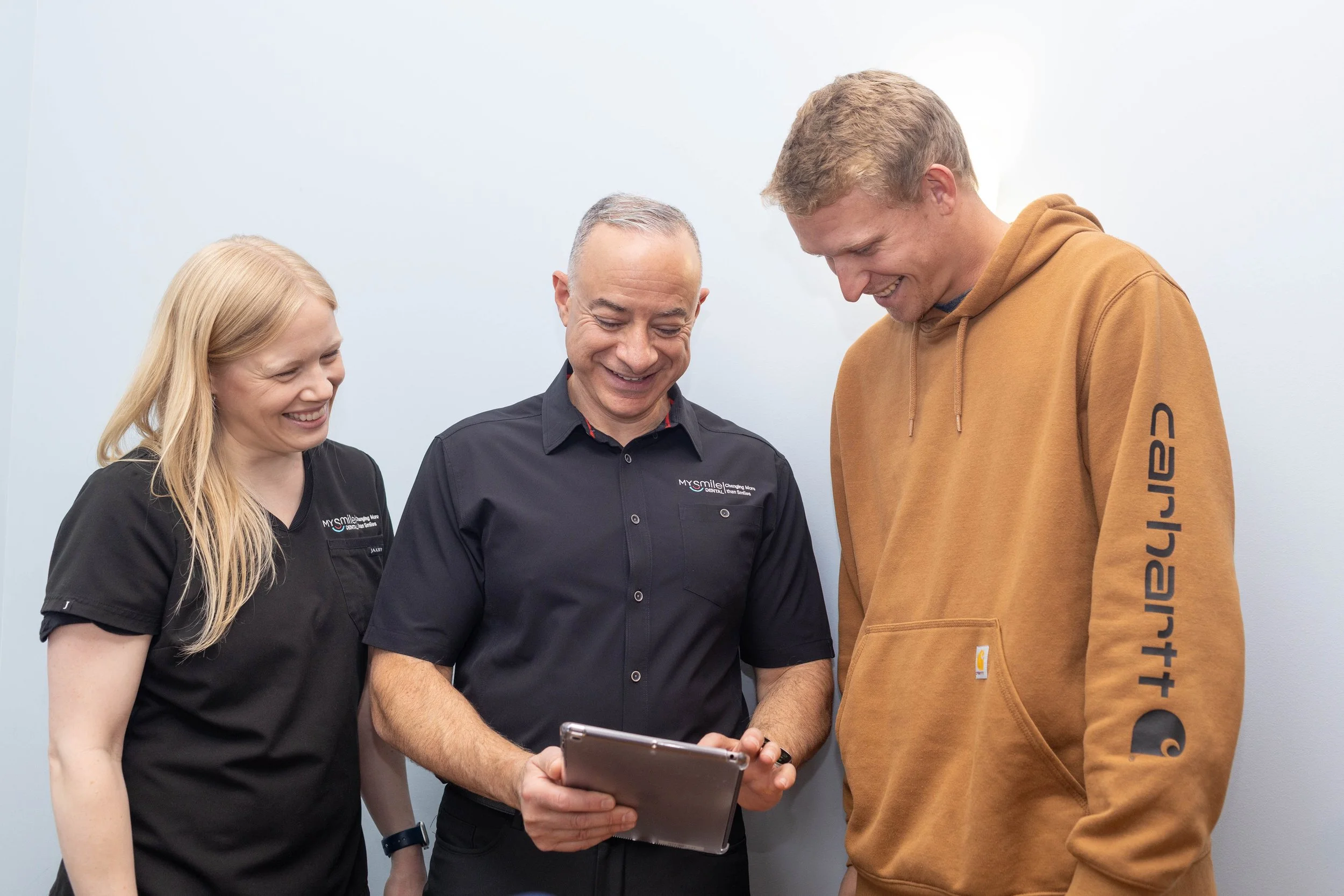 Three people, two women and one man, smiling and looking at a tablet together. The woman on the left has long blonde hair, is wearing a black shirt, and the man in the middle has short gray hair and is wearing a black shirt with a logo. The man on the right has short blonde hair and is wearing a brown hoodie with black lettering on the sleeve, all standing against a plain light gray background.