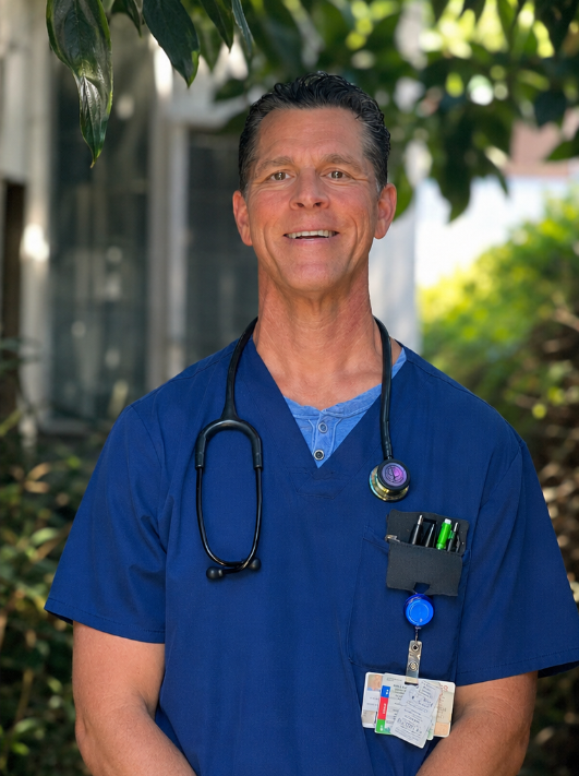 A smiling male healthcare professional wearing blue scrubs and a stethoscope outdoors, with trees and a building in the background.