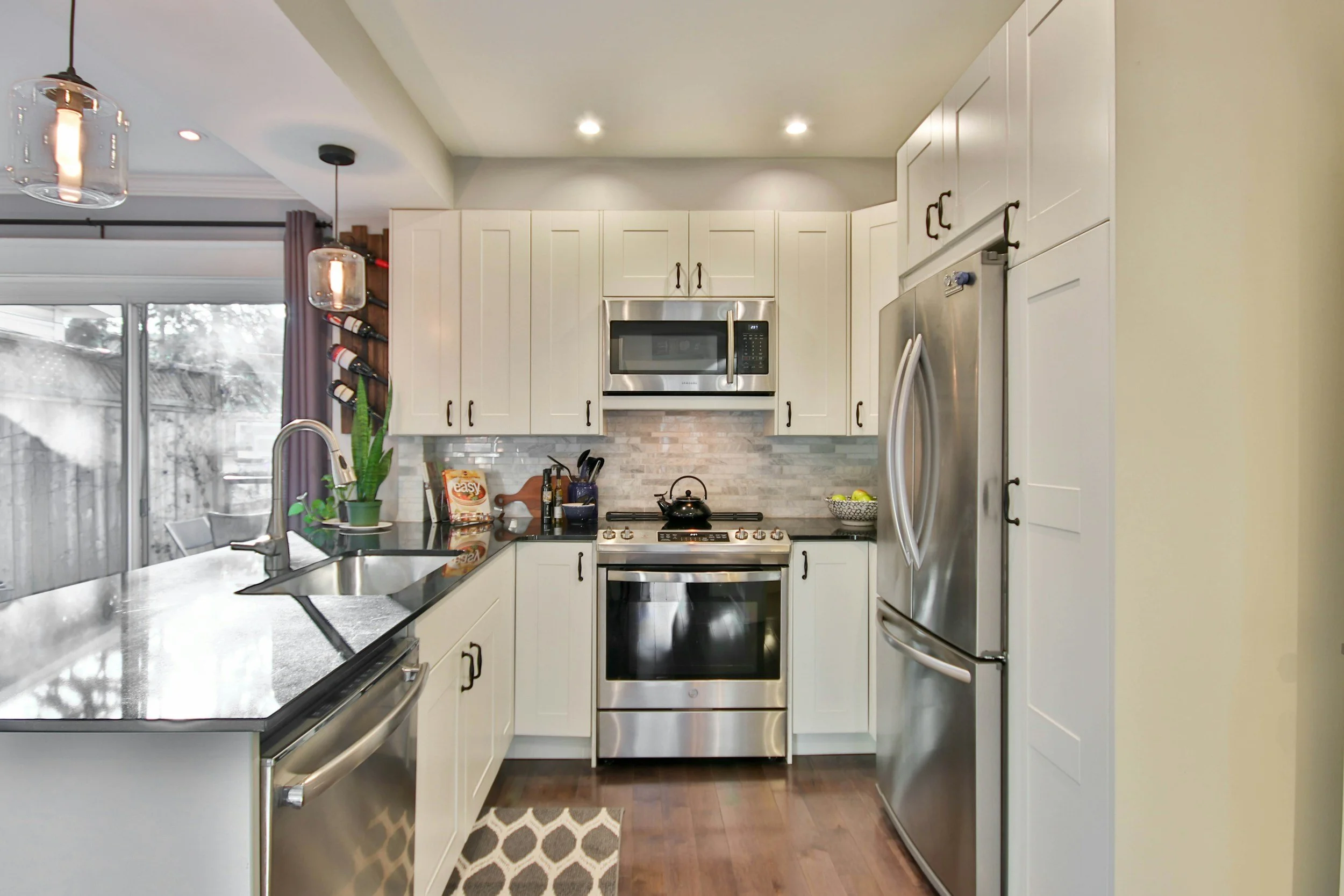 Modern kitchen with white cabinets, stainless steel appliances including a microwave, oven, and refrigerator, a black countertop with a sink, a window with purple curtains, pendant lights hanging from the ceiling, decorative plants, and a wine rack.