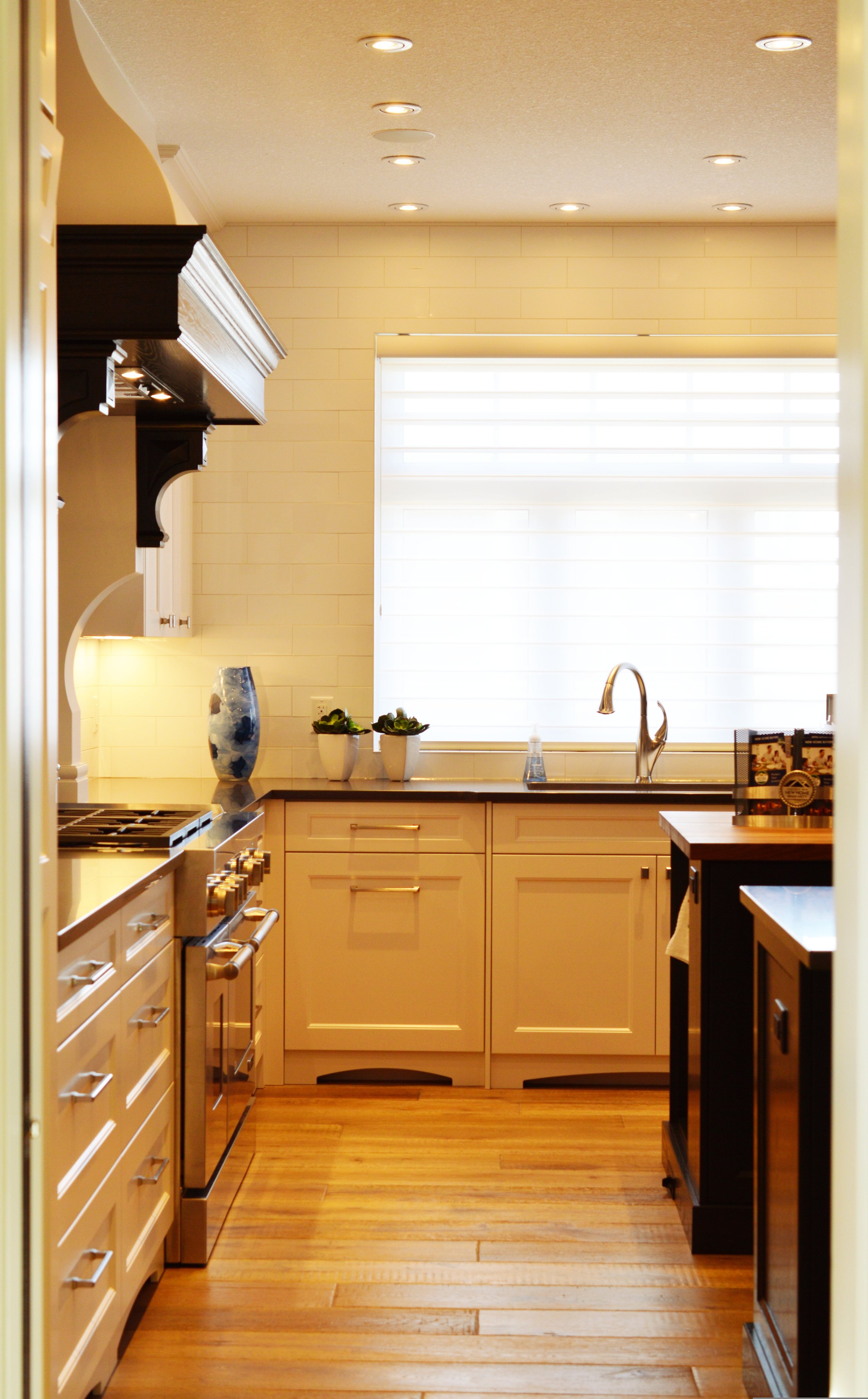 A kitchen with cream-colored cabinetry, a black kitchen island, wooden flooring, a window with white blinds, a sink with a chrome faucet, decorative vases and small plants on the counter, and a stove with a stainless steel finish.