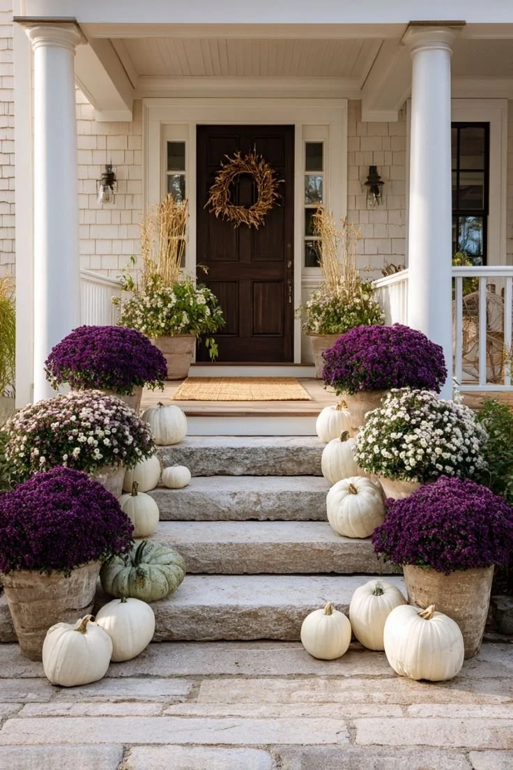 Front porch decorated with fall pumpkins, potted purple and white chrysanthemums, and a wreath on the door.