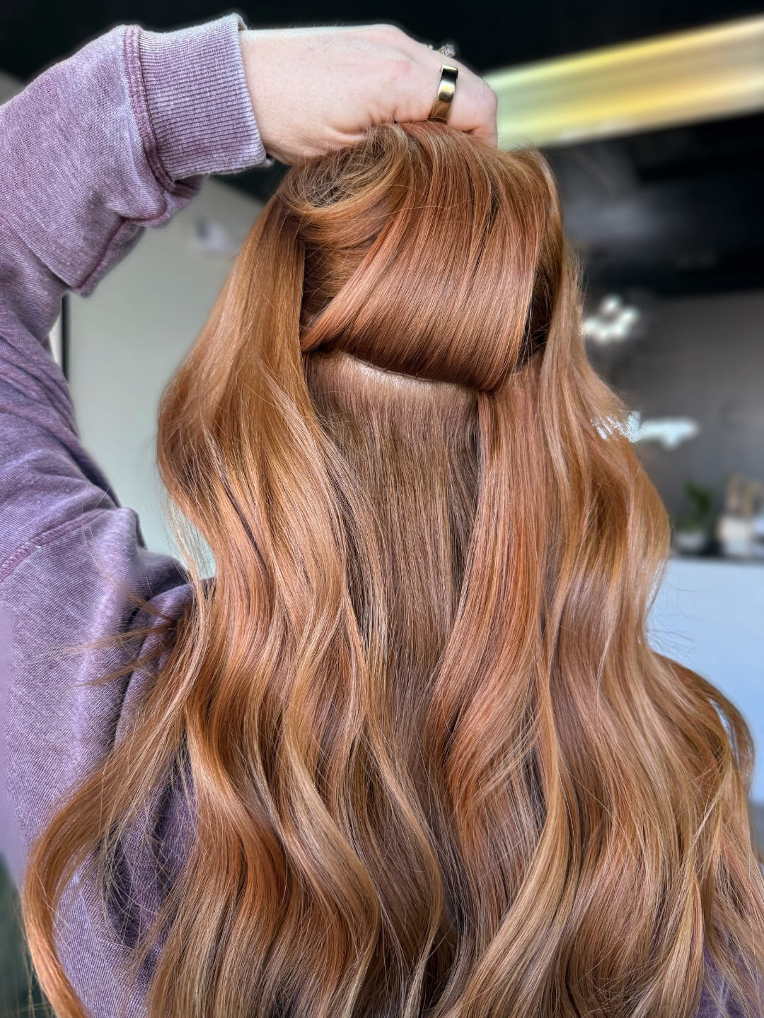A person with long, wavy, reddish-brown hair. They are holding their hair with their hand, showcasing the underside of their extensions. 