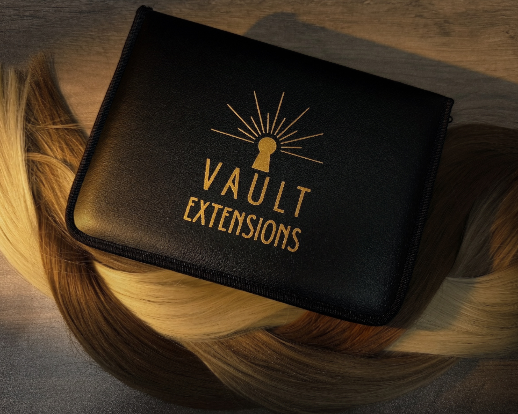 Close-up of a black hair extensions kit with gold text and design, placed on a wooden surface, surrounded by light brown and blonde hair extension wefts.