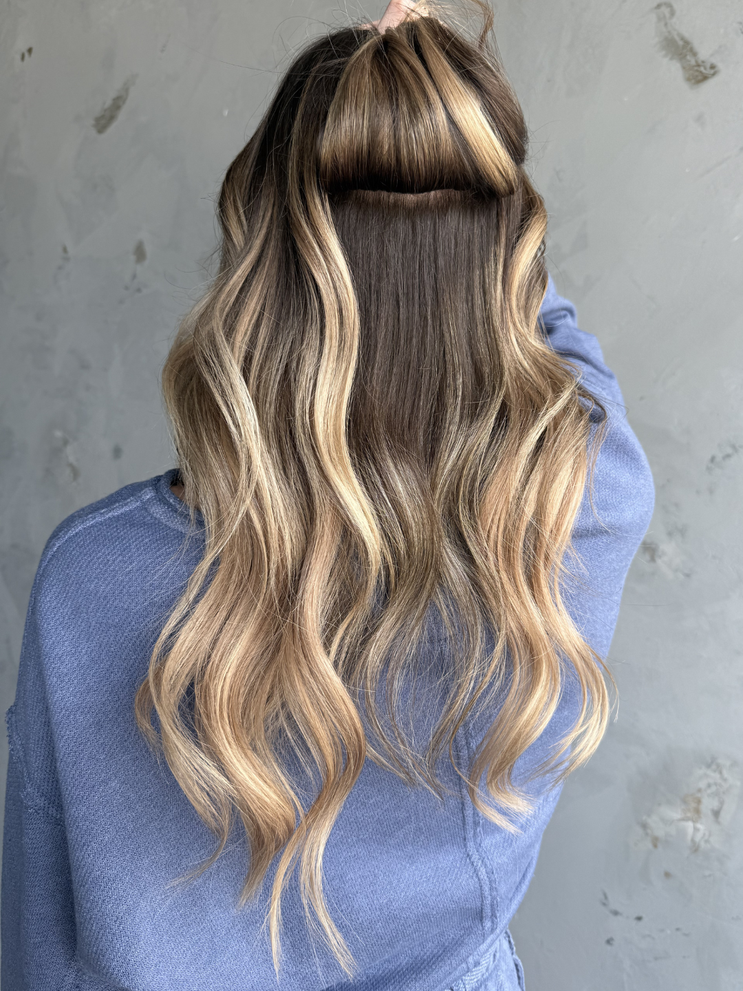 Back view of a person with long, wavy, blonde hair featuring darker roots, wearing a blue top, standing against a textured gray wall.