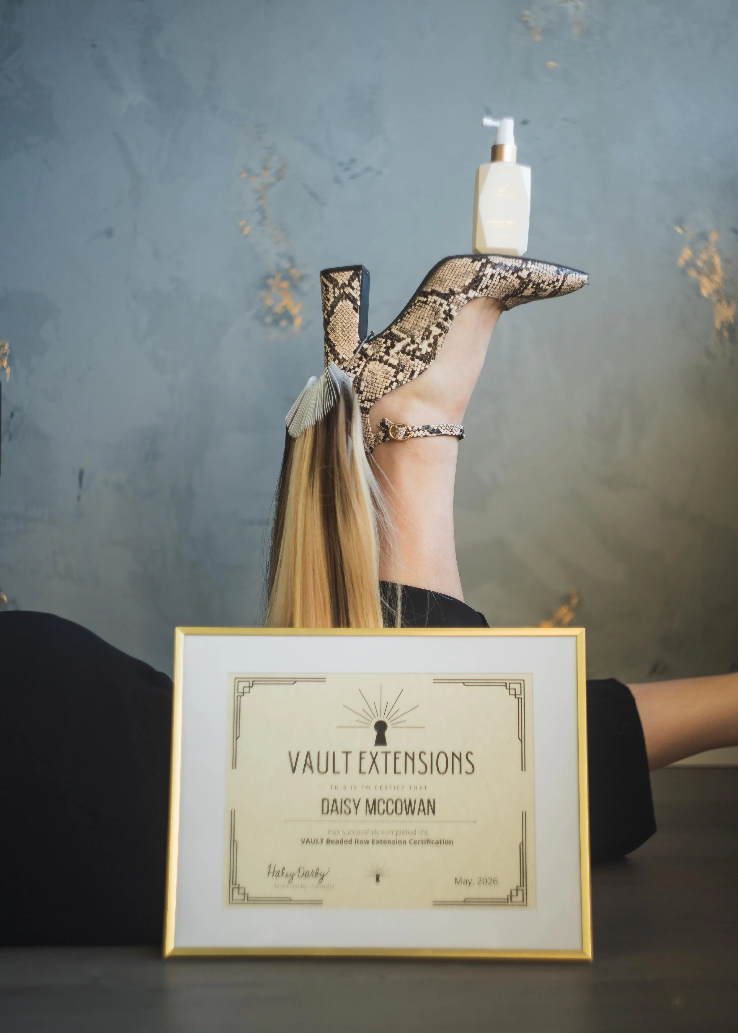 A person holding a certificate of completion for Vault Extensions, awarded in May 2026. The certificate is in a gold frame, and the person's hand is wearing a snake-print heel and an anklet. The background is a blue-gray wall with some gold accents.