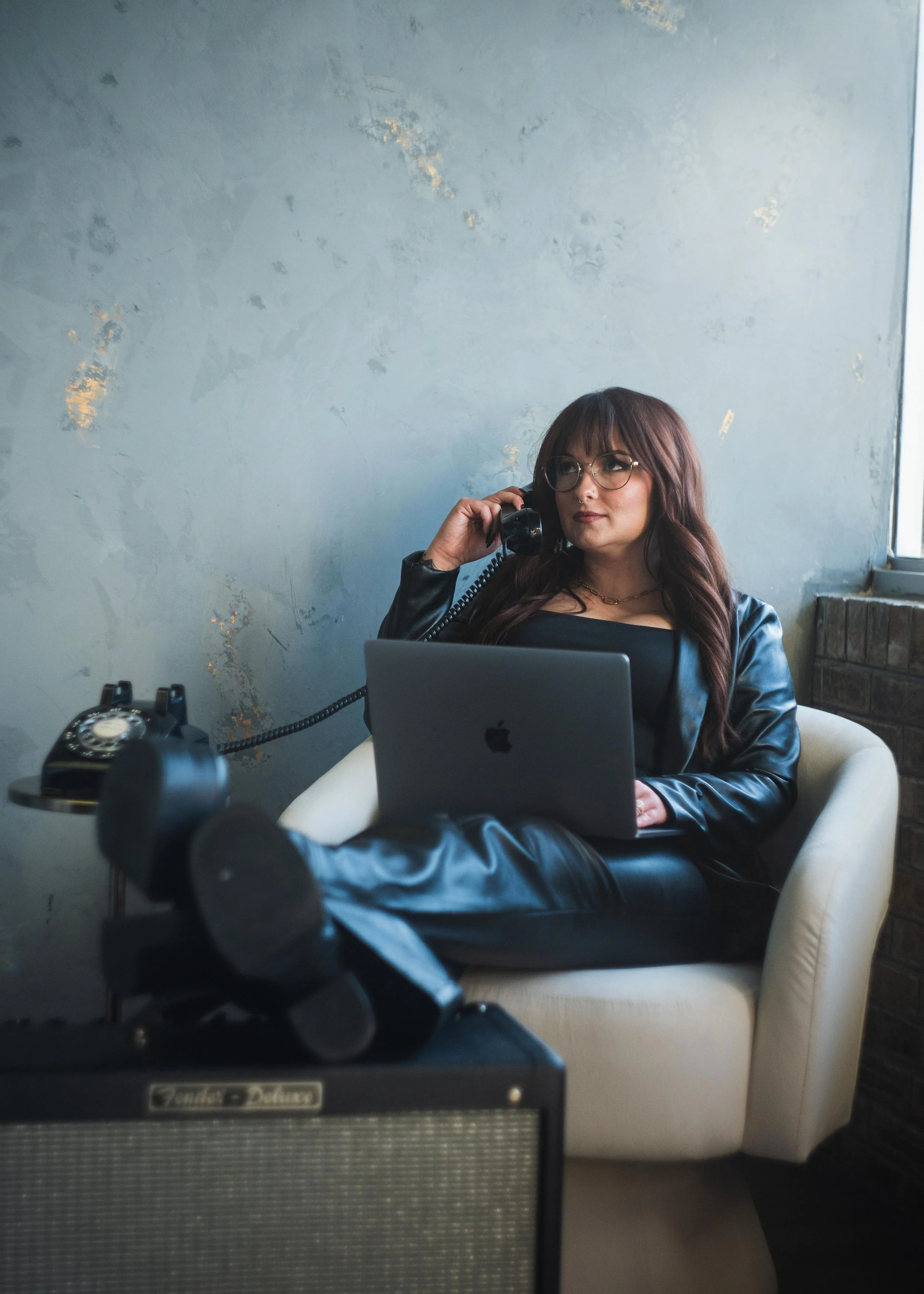 A woman with long brown hair, glasses, and a nose ring sitting in a cream armchair, talking on a black corded telephone while using a laptop. The scene is viewed through a mirror, with an antique radio visible in the foreground and a plain textured wall behind her.