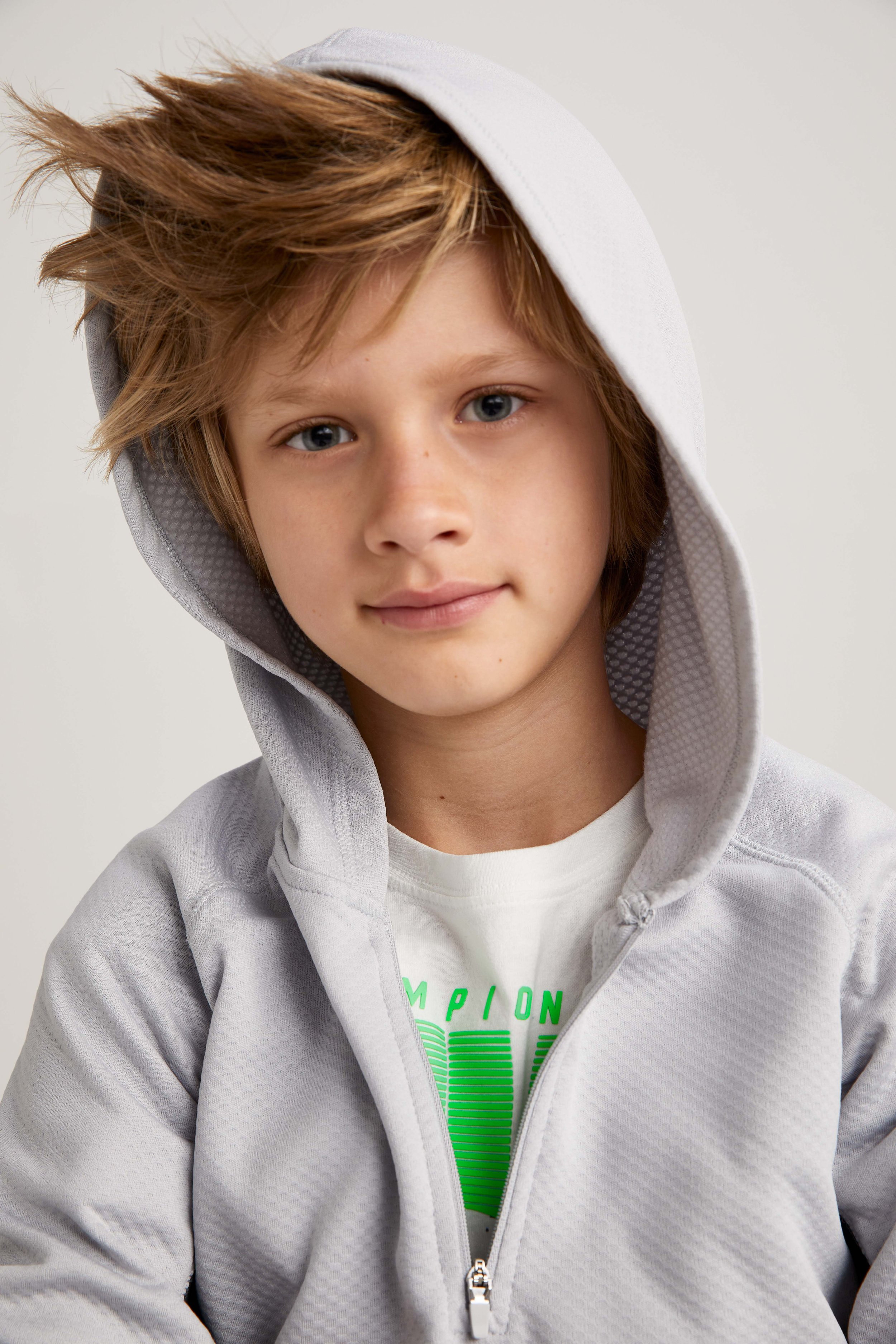 Close-up of a young boy with blue eyes and light brown hair, wearing a gray hoodie with the hood up and a white T-shirt with green text underneath.