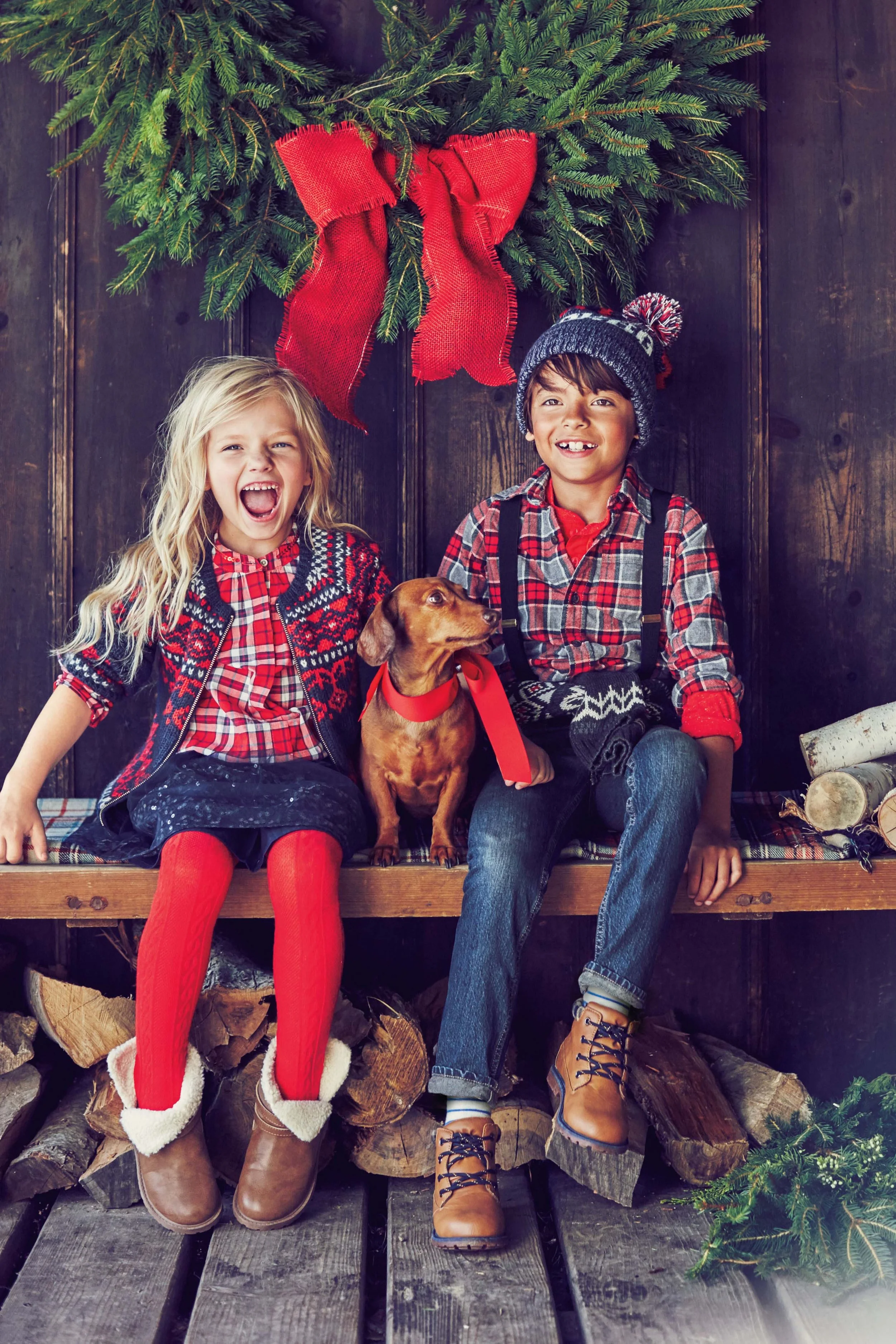 Two children, a girl and a boy, sitting on a wood log platform in front of a Christmas wreath with a large red bow, with a dog between them. The girl has long blonde hair, red tights, a plaid dress, and a dark vest, and is laughing. The boy has short