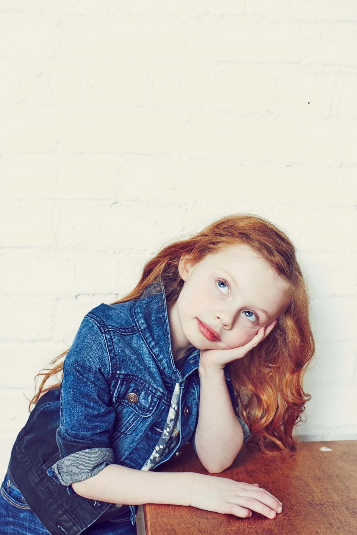 A young girl with long red hair and blue eyes resting her head on her hand, leaning on a wooden table, wearing a denim jacket, against a white brick wall background.