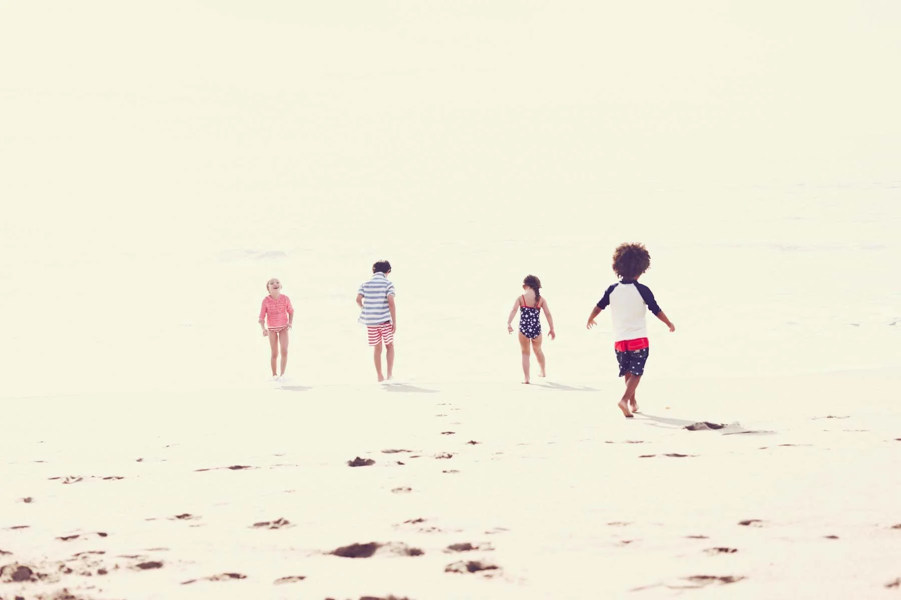 Five children walking on the beach, some in swimwear, with footprints in the sand and the ocean in the background.