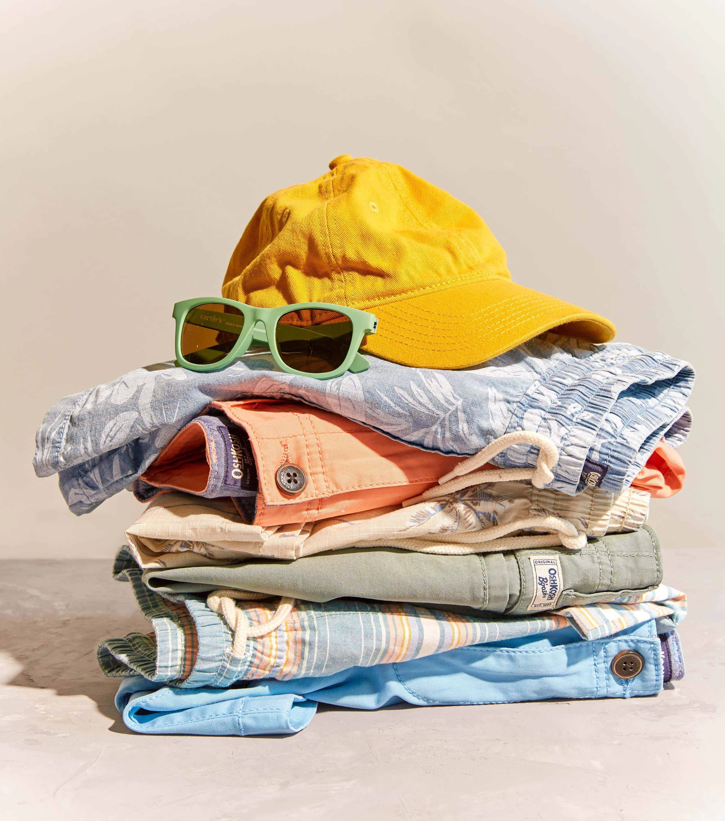 A stack of summer clothing, including shorts, a yellow baseball cap, and green sunglasses, on a concrete surface against a plain background.