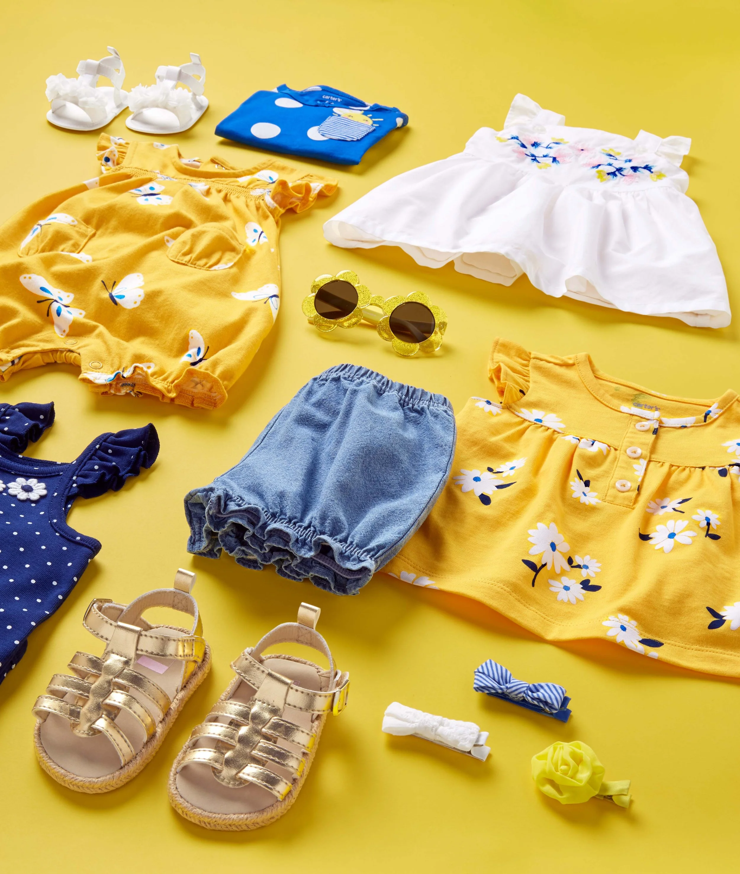 Children's clothing and accessories on a yellow background, including yellow butterfly-print dresses, white embroidered dresses, blue polka dot clothes, yellow sandals with straps, white sandals with flowers, sunglasses with flower-shaped gold frames