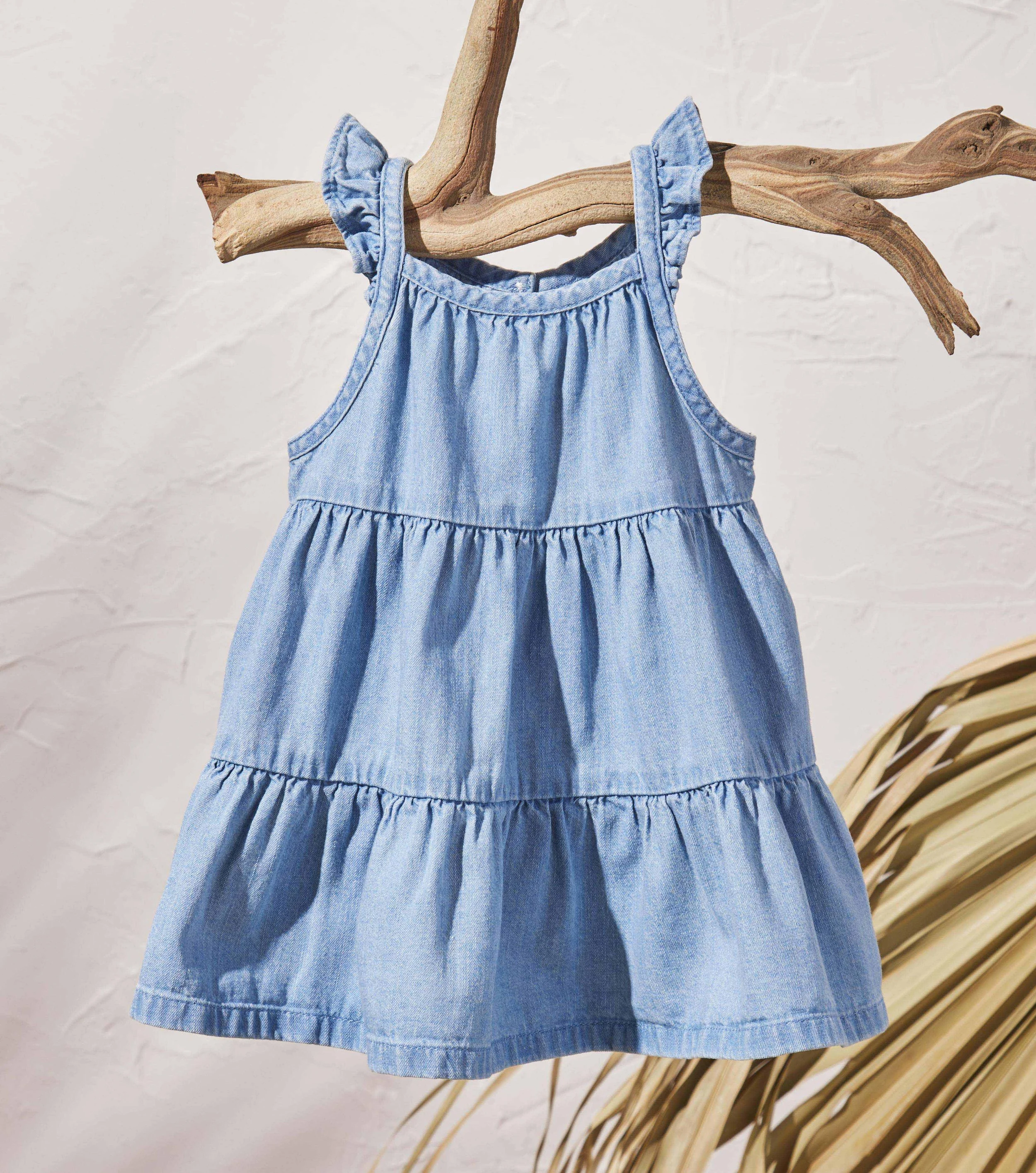 A light blue denim dress hanging on a natural wood branch, with a white textured wall in the background and dried palm leaves to the bottom right.