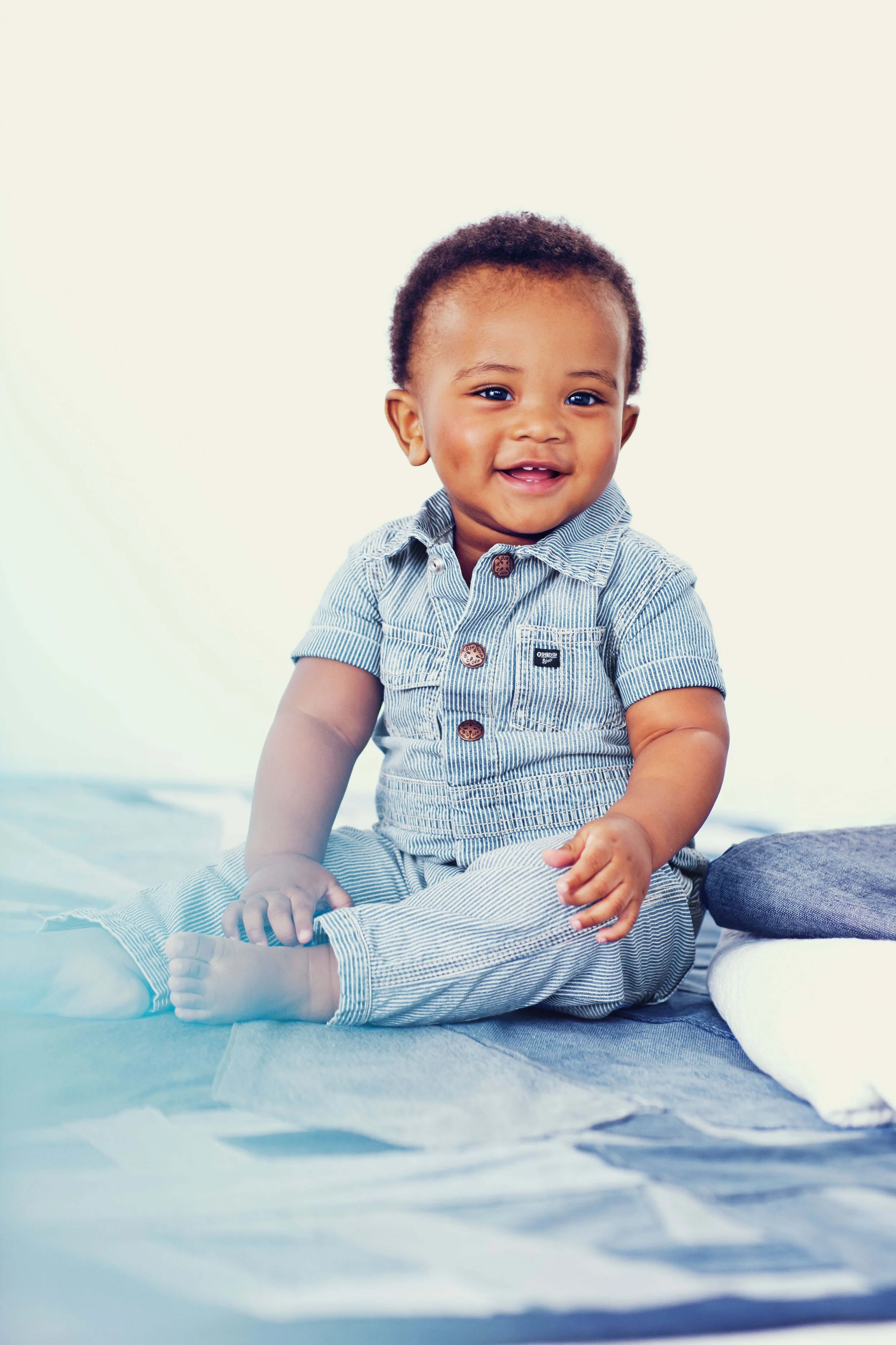 Smiling toddler with dark, curly hair and blue eyes, sitting on a blanket in a casual short-sleeve, striped button-up shirt. The background is plain white.