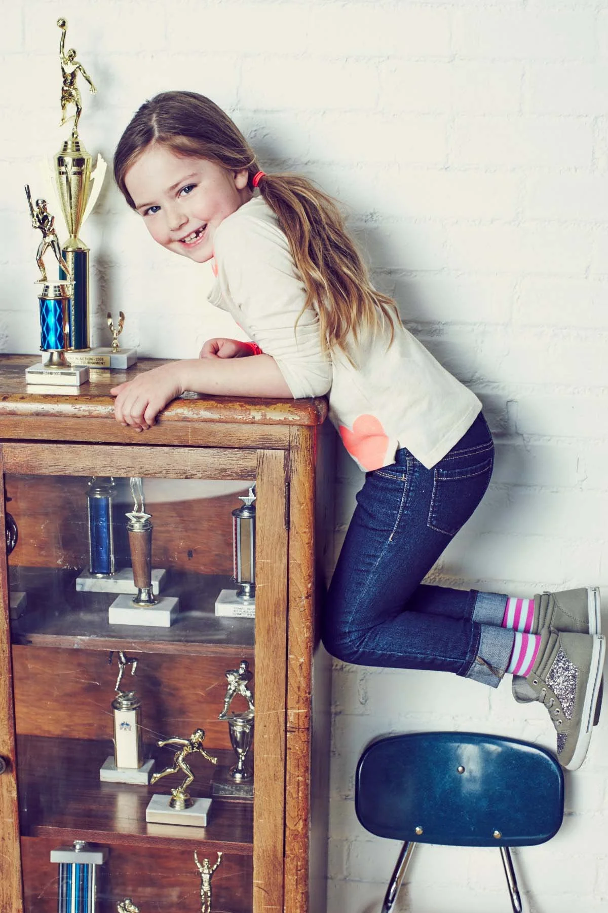 A young girl with long, light brown hair in pigtails, wearing a white shirt with orange patches on the sleeves, jeans, and glittery sneakers, is kneeling on a folding chair next to a wooden display case of trophies, smiling at the camera.