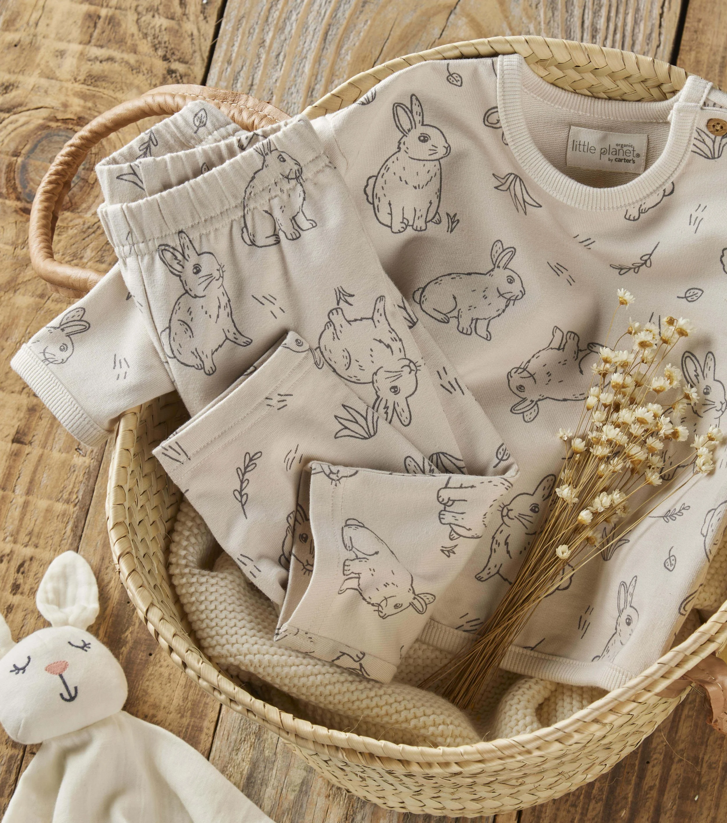 Baby clothes with bunny prints in a woven basket, on a wooden surface, with a plush animal toy and some dried flowers nearby.