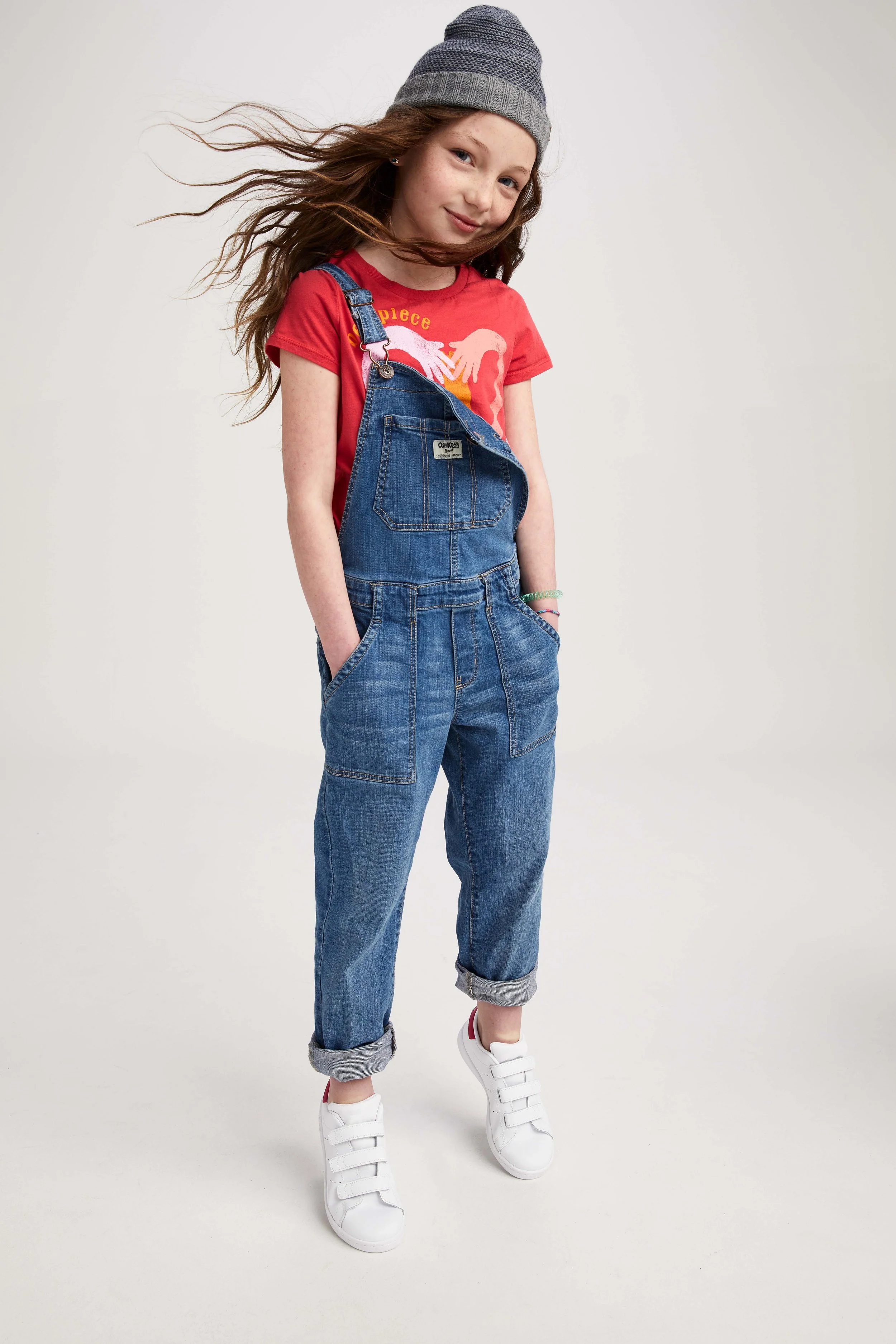 A young girl with long hair wearing a gray knit beanie, red t-shirt, and blue denim overalls with rolled-up cuffs, white sneakers, and colorful bracelets, standing against a plain white background.