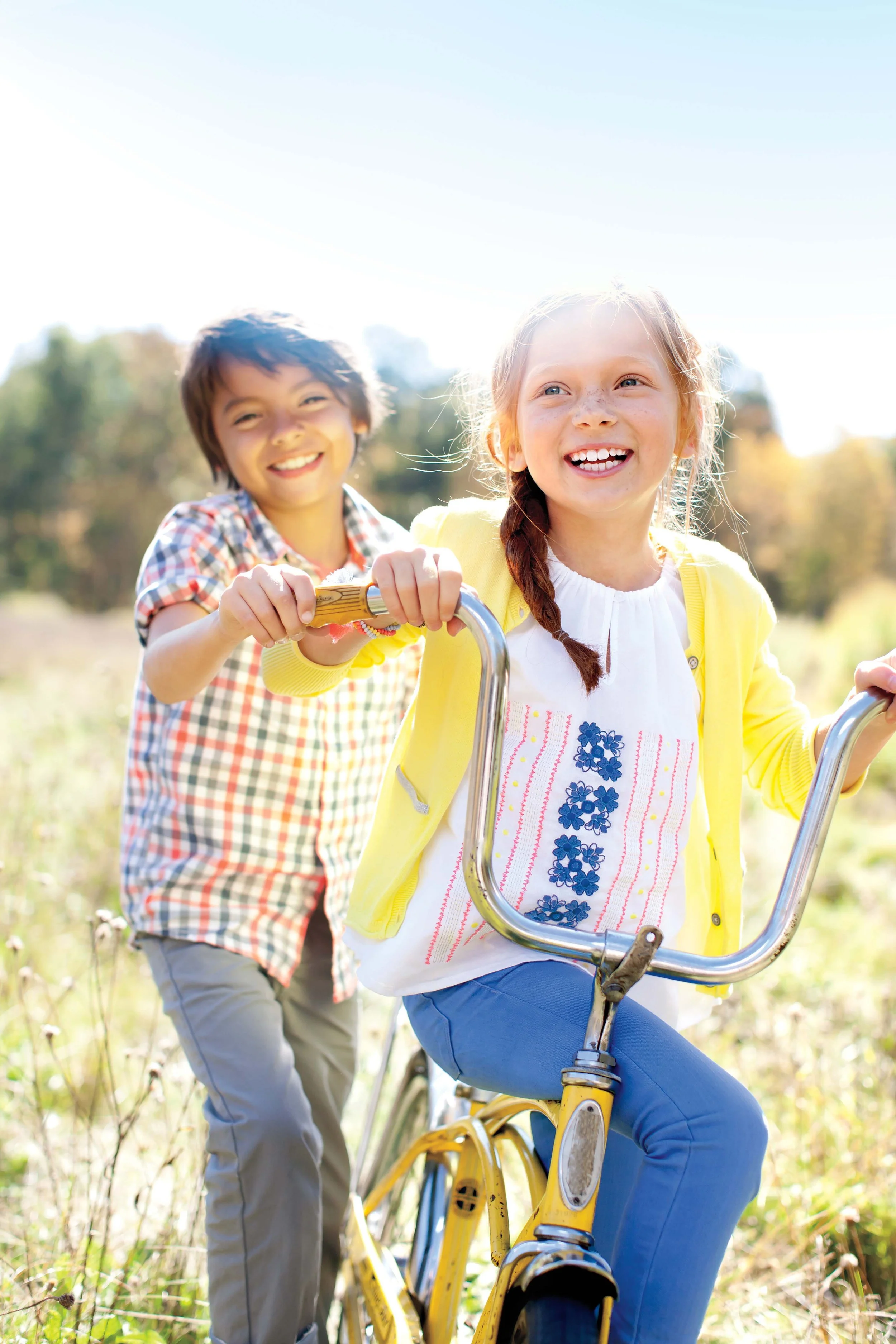 Two young girls outdoors, one riding a yellow bicycle and the other walking behind her, both smiling and enjoying a sunny day in a natural setting with greenery.